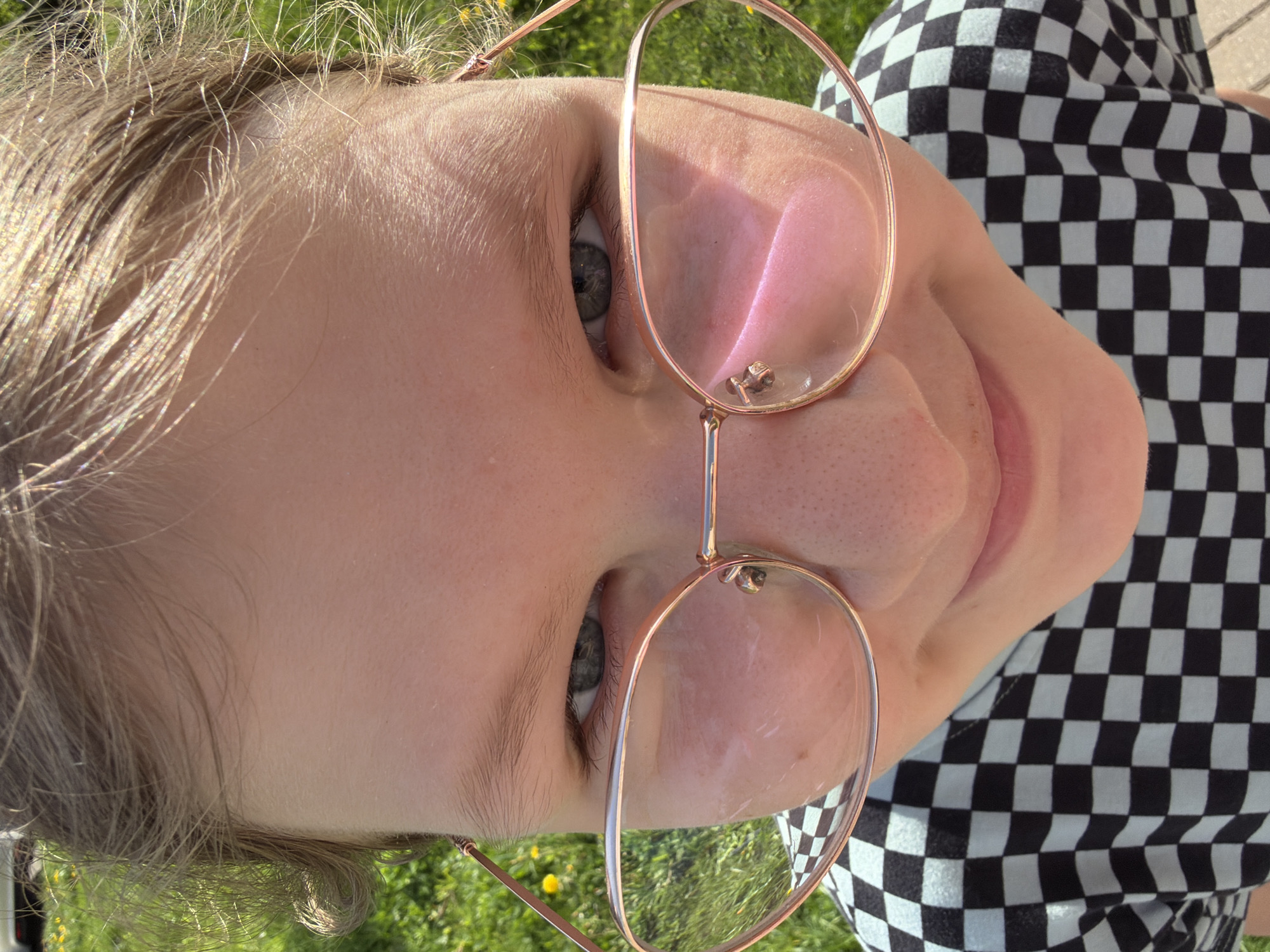 A close-up, ground-level perspective photograph shows a person with light skin and light-colored hair, wearing gold-rimmed glasses. The subject is tilted in the frame, with their eyes visible and gazing toward the camera. Their nose is prominent in the center of the frame due to the angle. A slight smile can be seen. The person wears a black and white checkerboard pattern shirt. The background consists of green grass with scattered small yellow flowers, indicating an outdoor setting. The specific location is not evident from the image.