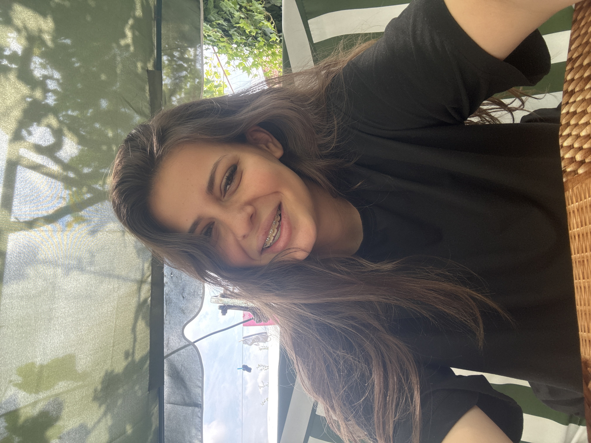The media file shows a portrait of a young woman with long, brown hair, smiling at the camera. She is wearing a black t-shirt and has braces on her teeth. The image appears to be taken outdoors on a sunny day. In the background, there is greenery, possibly trees or bushes, and a blue sky with some clouds. There are also some striped green and white fabric elements visible, which could be part of an awning or tent. A woven texture, possibly from furniture, is visible on the right side of the frame. The overall impression is a casual and relaxed moment, perhaps taken during leisure time. The lighting suggests it is daytime, likely late morning or afternoon. There is no visible text in the image. The location context is Govedare, Bulgaria.
