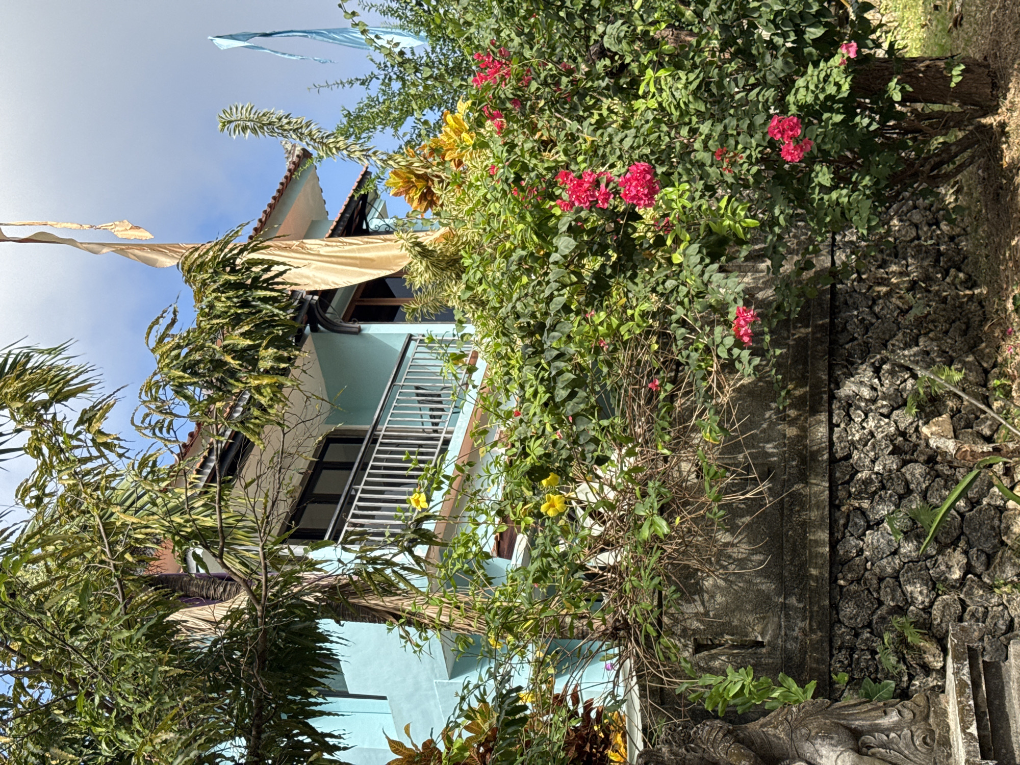 In Kuta Selatan, Indonesia, a light blue building with a white railing on a balcony is partially obscured by lush green foliage and vibrant pink and yellow flowers. A stone wall, constructed from rough, dark stones, runs along the bottom of the frame, topped by a section of rough-textured concrete. To the left, a carved stone statue is visible. The sky above is a bright blue with scattered clouds, and two fabric banners, one gold and one blue, are seen hanging from poles. The scene depicts a residential building surrounded by a tropical garden.