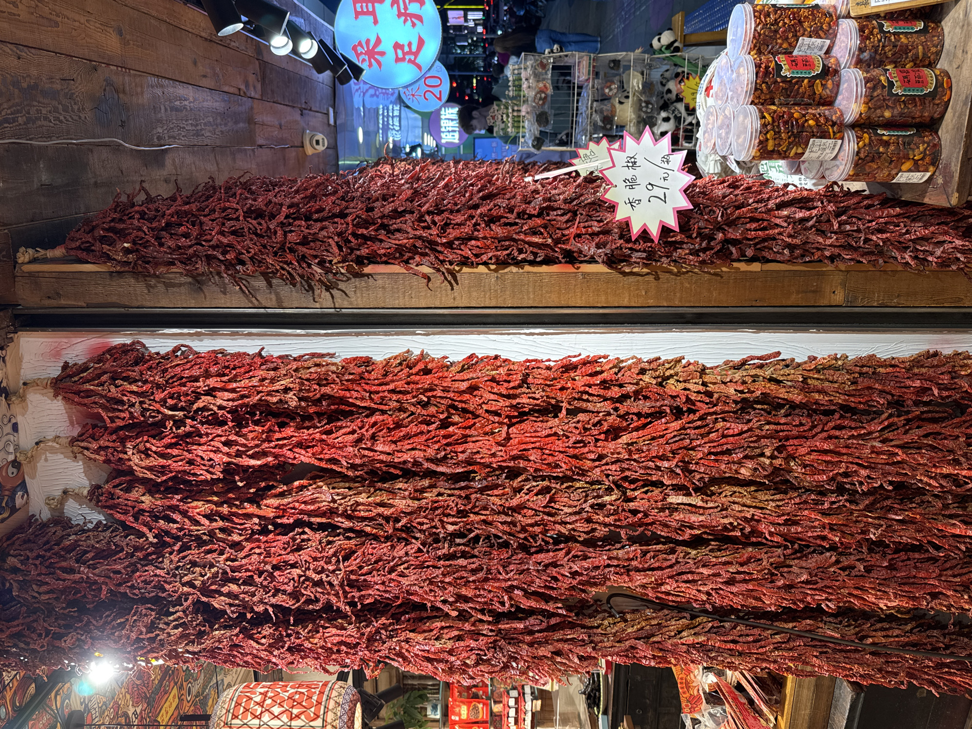The image depicts a display of dried red peppers in the Jinjiang District of China. The peppers are tied in bundles and hung from a ceiling and placed on a wooden shelf. A sign displays a price of 29 yuan. Additional food products are visible in jars on the shelf. The shop appears to be located on a busy street.