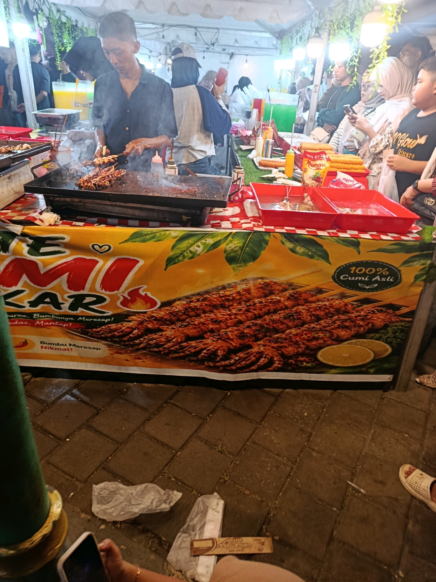 In Balaraja, Indonesia, a street food stall is pictured at night. A vendor, wearing a dark blue shirt, is grilling skewers of calamari over a flat-top grill, with visible smoke rising. The stall is decorated with a banner featuring a large image of grilled calamari and text in Indonesian, including "CUMI ASLI" (Authentic Calamari) and promotional phrases like "Gurih, Bumbunya Meresap!" (Savory, the spices penetrate!). Several people, including women wearing hijabs, are visible in the background, some looking at their phones or browsing the food. The stall has red trays holding food items and bottles of condiments. The ground is paved with interlocking bricks. In the foreground, a green pole is visible, and a person's hand holding a smartphone is in the lower left corner. Discarded paper wrappers are on the ground near the stall. The overall scene depicts a lively night market atmosphere with a focus on grilled seafood.