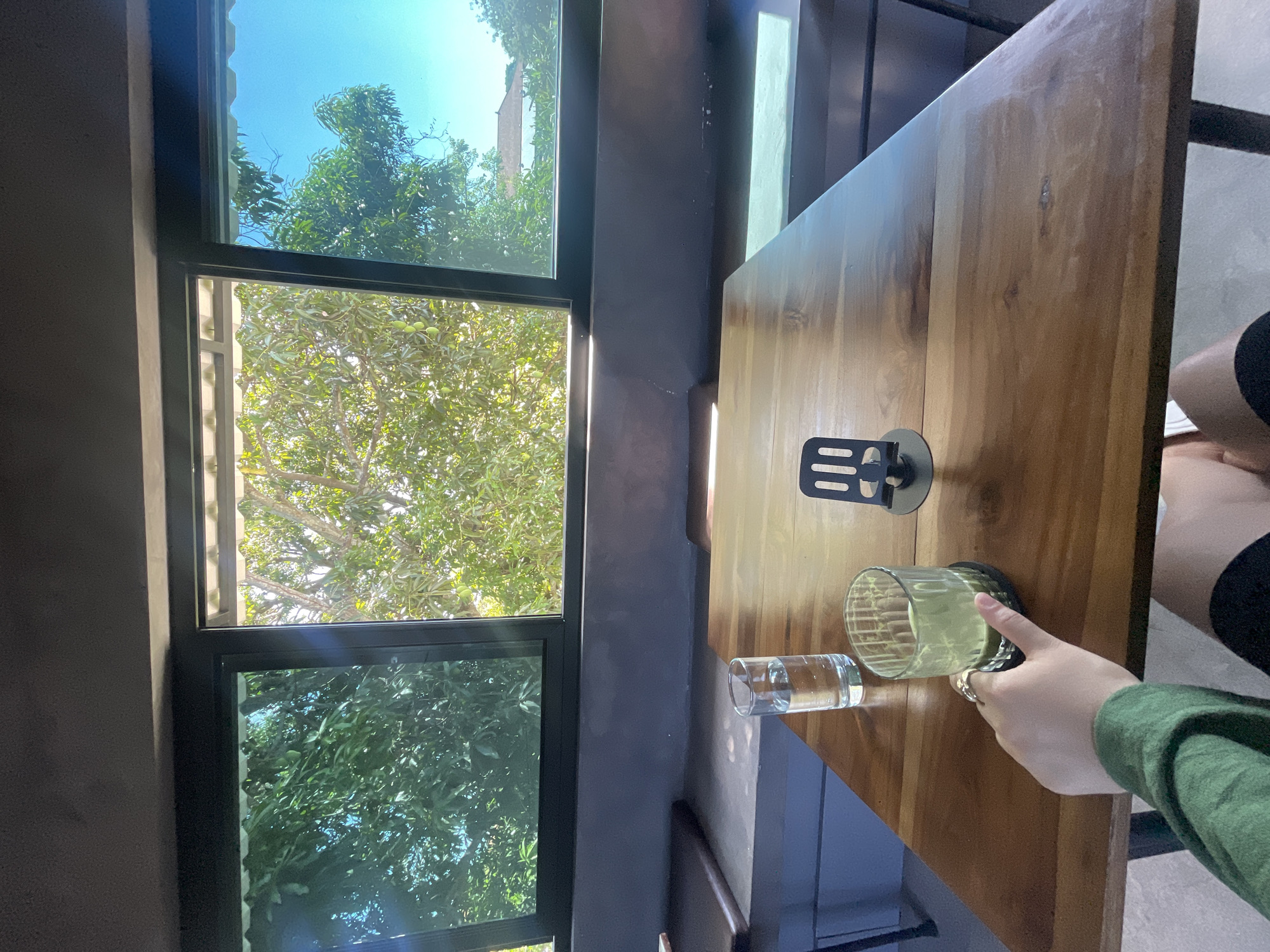The image depicts a close-up view of a wooden table in an indoor setting, likely a cafe or restaurant, with a large window offering a view of lush green trees and a bright blue sky. On the table, there are two glasses, one filled with clear liquid and another with a green beverage, possibly matcha. A person's hand, adorned with a ring, is reaching out towards the glass of green drink. The scene is illuminated by natural light filtering through the window, creating subtle shadows on the table. The overall atmosphere is serene and relaxed. The location is Kuta Selatan, Indonesia, a region known for its beautiful beaches and vibrant cafes.