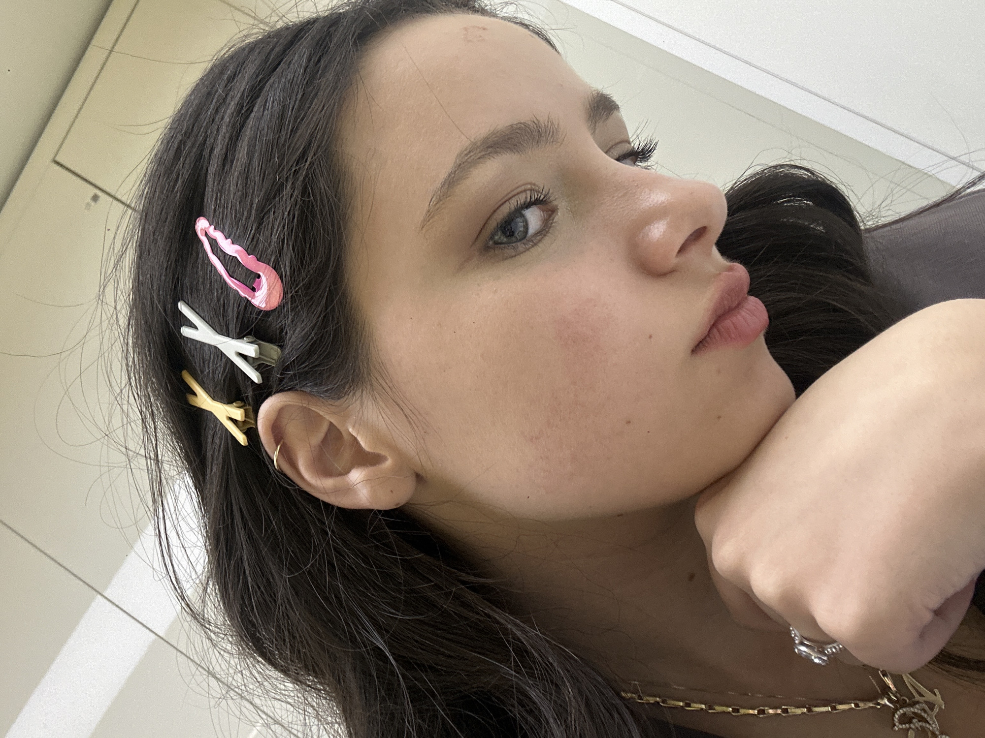 A close-up, slightly angled headshot shows a young woman with long, dark hair. She has fair skin, light blue eyes, and full lips. Three decorative hair clips adorn the left side of her hair: one pink, one white, and one yellow. She is wearing a small gold hoop earring. Her face rests on her hand, which is adorned with a silver ring with a stone. A gold chain necklace is visible around her neck. The background is a blurred, light-colored wall, and no indication is given about a city or country.