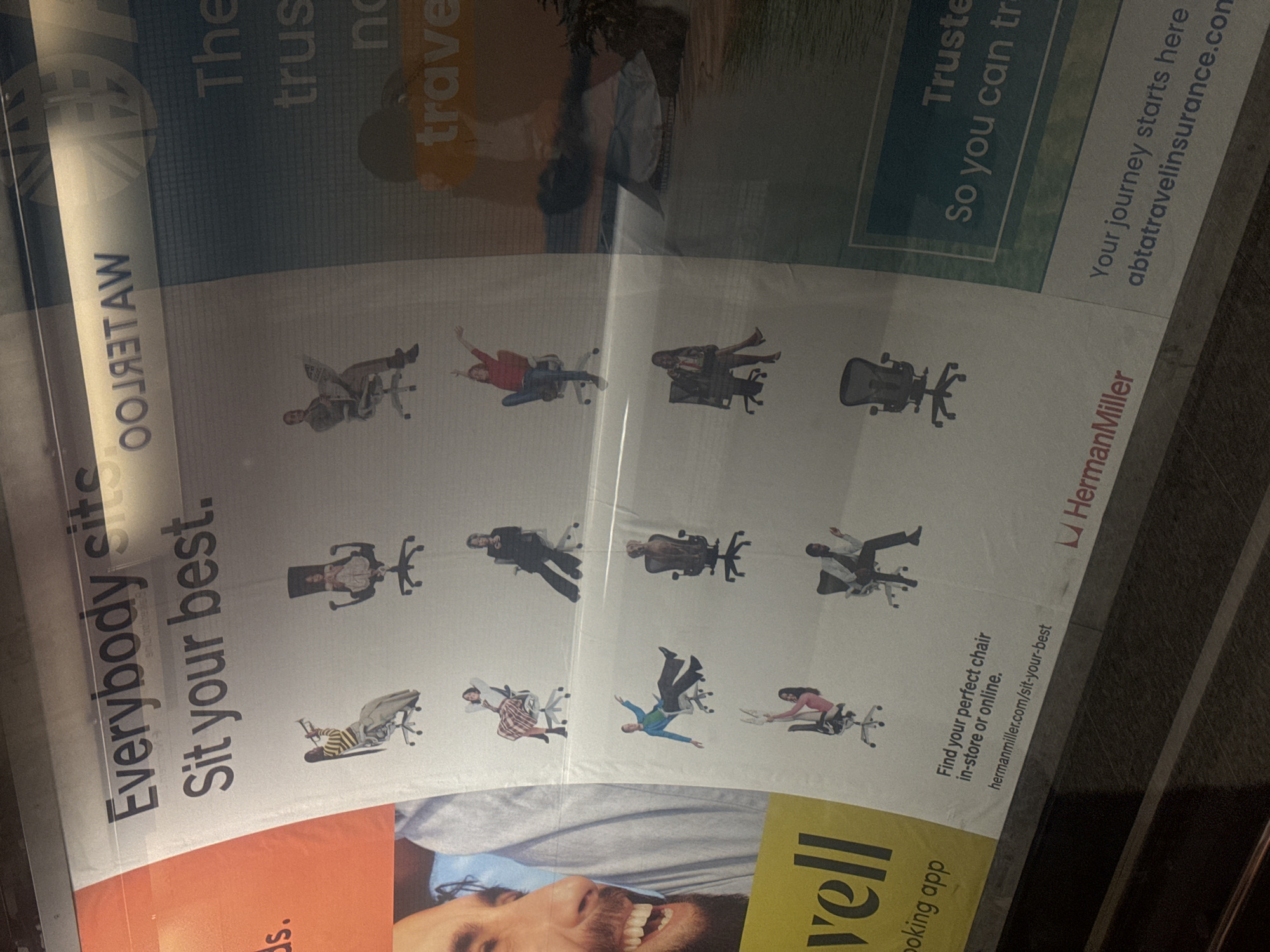 The image shows an advertisement for Herman Miller office chairs, prominently displaying the tagline "Everybody sit. Sit your best.". The advertisement features various people sitting in different styles of office chairs, set against a predominantly white background. Text elements include "Find your perfect chair in-store or online" and the Herman Miller website. Adjacent to the ad is another advertisement, visible partially, that contains the word "travel." The photo appears to have been taken at the Waterloo Station, in the city of London, United Kingdom. The image is taken at an angle.