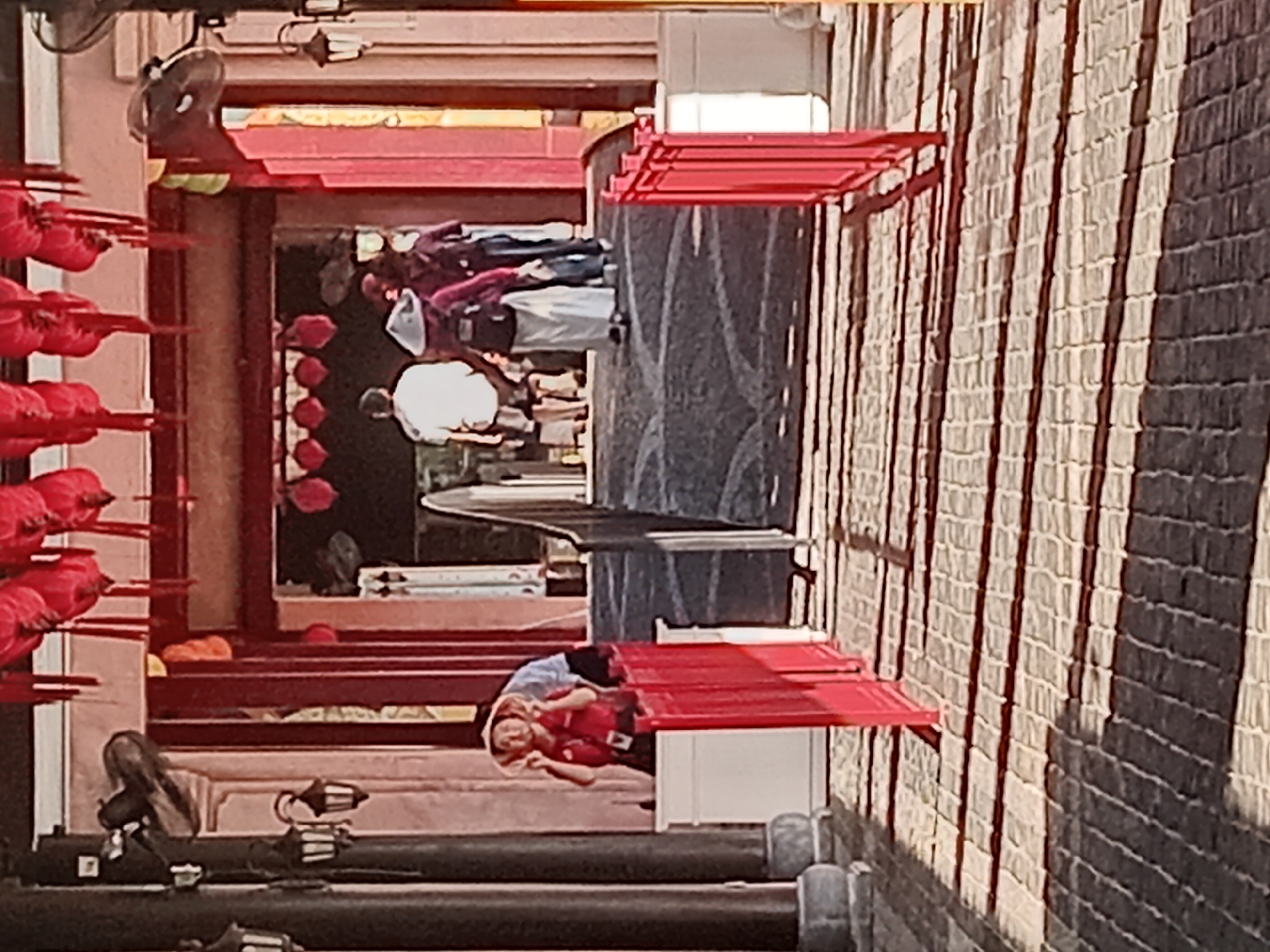 The image captures a scene within a culturally inspired architectural setting, likely a tourist attraction or themed area in Đà Nẵng, Vietnam. In the foreground, a red fence directs the viewer's eye towards a pathway lined with stone or brickwork casting striped shadows from overhead structures, suggesting it is daytime and the sun is at an angle.
To the left, a person wearing a red shirt and a straw hat is partially visible behind a white barrier, looking towards the viewer with their hands near their face. This individual appears to be an employee or performer, given their attire.
The background reveals an arched passageway adorned with numerous red lanterns, indicative of Asian festivals or decor. Several people are seen walking through this archway. One person in the center is wearing a traditional conical hat and a long white skirt, carrying a backpack. To their right, another individual in a maroon shirt and jeans is walking away from the camera. Further back, a person in a white shirt and shorts is walking towards the archway.
The architecture features dark columns and red accents, contributing to a traditional aesthetic. The overall atmosphere suggests a vibrant and possibly historical or entertainment-focused location.