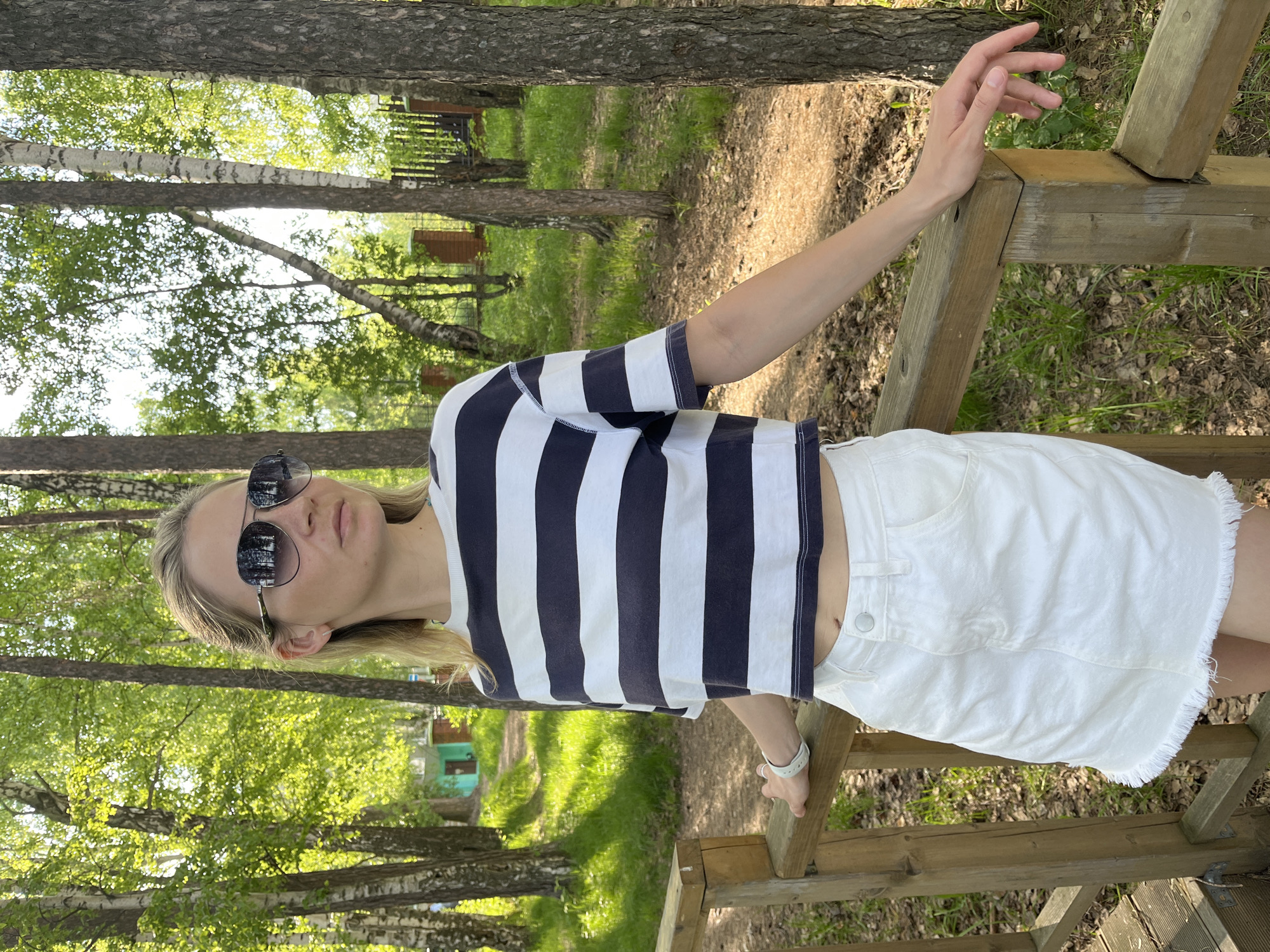A woman stands outdoors on a wooden structure, likely a bridge or raised platform, in a wooded area. She wears a navy and white striped cropped t-shirt, a white denim skirt, and sunglasses. Her left hand rests on a wooden railing while her right arm extends towards another part of the structure. The background consists of trees, including birch trees, and patches of green grass. The photo appears to be taken in a natural setting. Based on the request to determine the location within Chaikovsky, Russia, and considering the presence of birch trees and wooded environment, it is likely that this photo was taken at a public park or a recreational area near the river Kama, where such natural elements are prevalent.