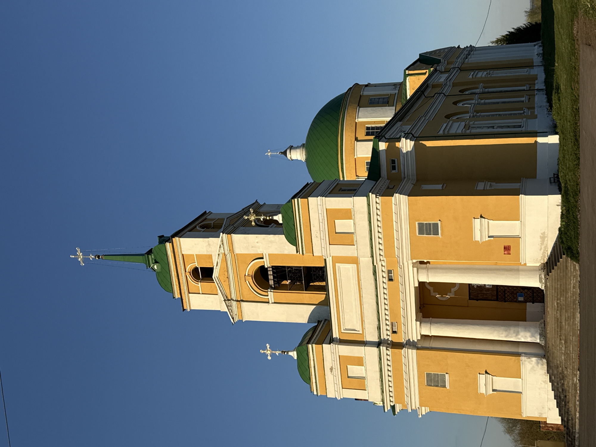 The image shows the Church of the Archangel Michael in Мазунино, Russia. It is a yellow and white building with green domes and crosses on top. The sky is blue. The church has a bell tower. There are steps leading up to the entrance.