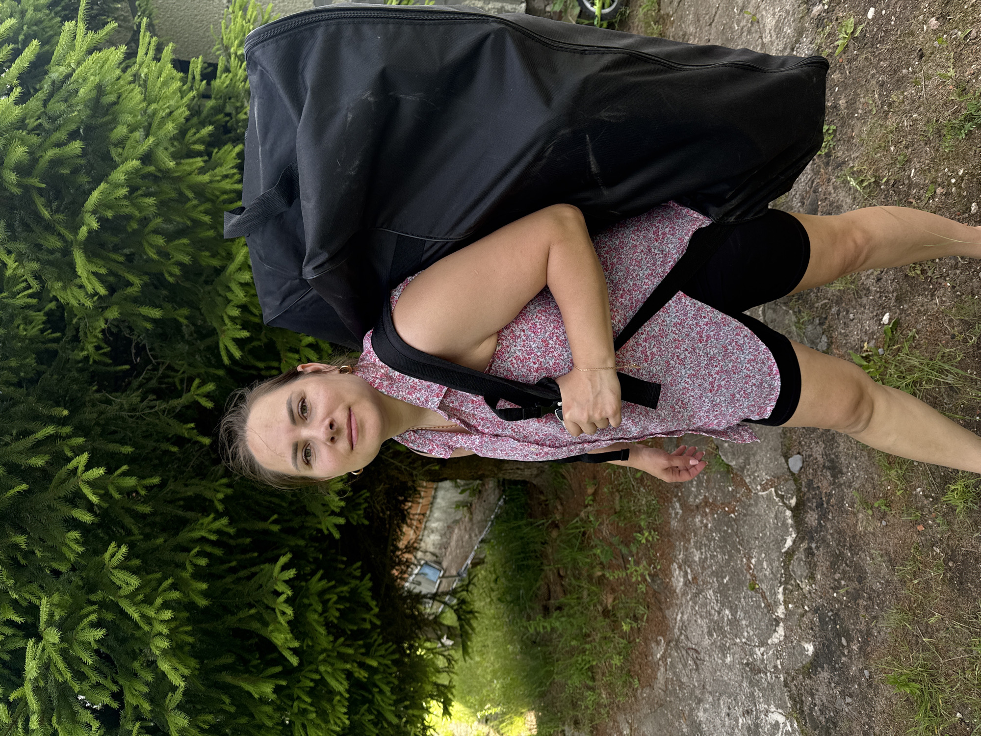 A woman is shown from the chest down, carrying a large black bag over her left shoulder. She is wearing a short-sleeved pink dress with a floral pattern and black shorts. Her right arm is bent at the elbow and her hand is resting on the bag. Her left hand is also on the bag. She is looking at the camera with a neutral expression. The bag appears to be a duffel bag or travel bag. The scene is outdoors, with lush green trees and foliage visible on the left side of the image. A gray concrete path or ground is visible on the right. The lighting suggests it is daytime, possibly late afternoon. The overall setting appears to be a backyard or a rural area. No text is visible in the image.