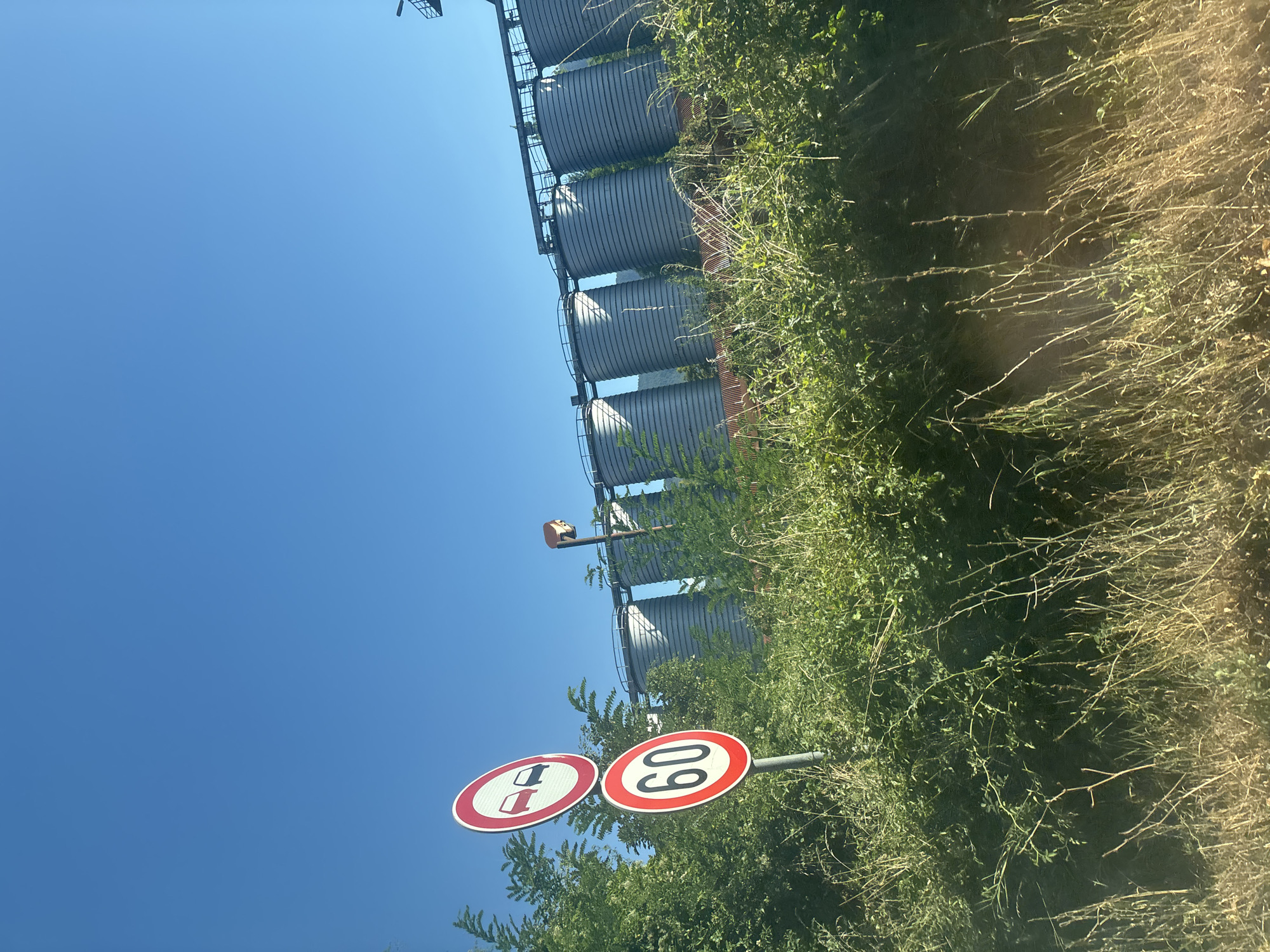 The image shows a road sign with two circular signs. The top sign depicts two cars, one red and one black, with a red circle and a red diagonal line through them, indicating "no overtaking". The bottom sign has a red circle with the number "60" inside, indicating a speed limit of 60 kilometers per hour. These signs are mounted on a metal pole and are situated among lush green foliage and dry grass. In the background, a row of tall, cylindrical silos, likely part of an industrial facility, are visible against a clear blue sky. The silos are metallic and appear to be weathered. The overall scene suggests an outdoor setting, possibly on the outskirts of a town or near an industrial area. The presence of traffic signs implies a roadway is nearby. This scene is located in the city of Pravets, Bulgaria.