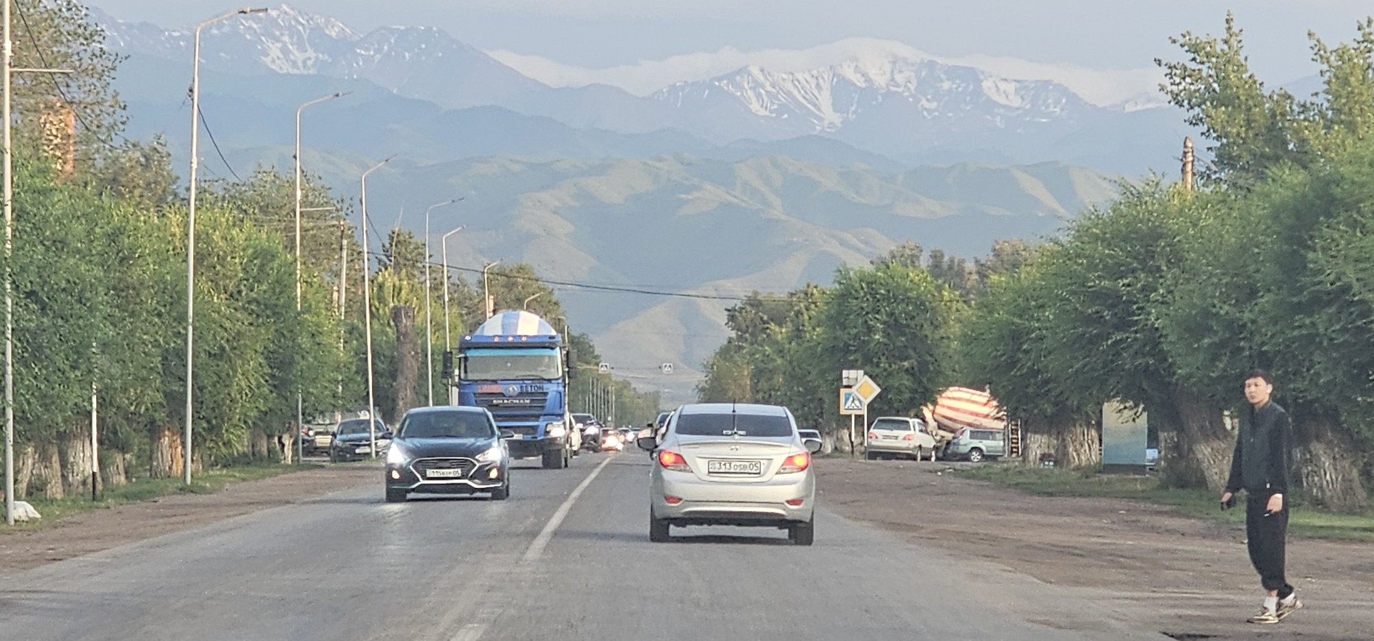 The media file depicts a street scene in Жамбыл, Kazakhstan, likely during the late afternoon or early evening, given the soft, diffused lighting and long shadows. The setting is a paved road with multiple lanes, bordered by lush green trees on both sides. In the background, majestic, snow-capped mountains rise against a hazy, pale blue sky, suggesting a rural or mountainous region.
Several vehicles are on the road, including a blue truck with a distinctive white and blue striped cab and a white sedan in the foreground, which has a license plate with the text "313 OSP 05". Other cars are visible further down the road, indicating moderate traffic. On the right side of the road, a young man is walking, dressed in dark clothing and what appear to be athletic shoes. He is holding something in his right hand, possibly a cigarette.
The scene conveys a sense of everyday life in a scenic location. The presence of trees and mountains suggests an area with natural beauty, while the road and vehicles indicate a populated area. The weather appears to be clear and mild. The overall mood is calm and ordinary, a snapshot of a moment in time on a road leading towards or away from the imposing mountain range. The general atmosphere is one of transition, with the road acting as a conduit between human activity and the grandeur of nature.