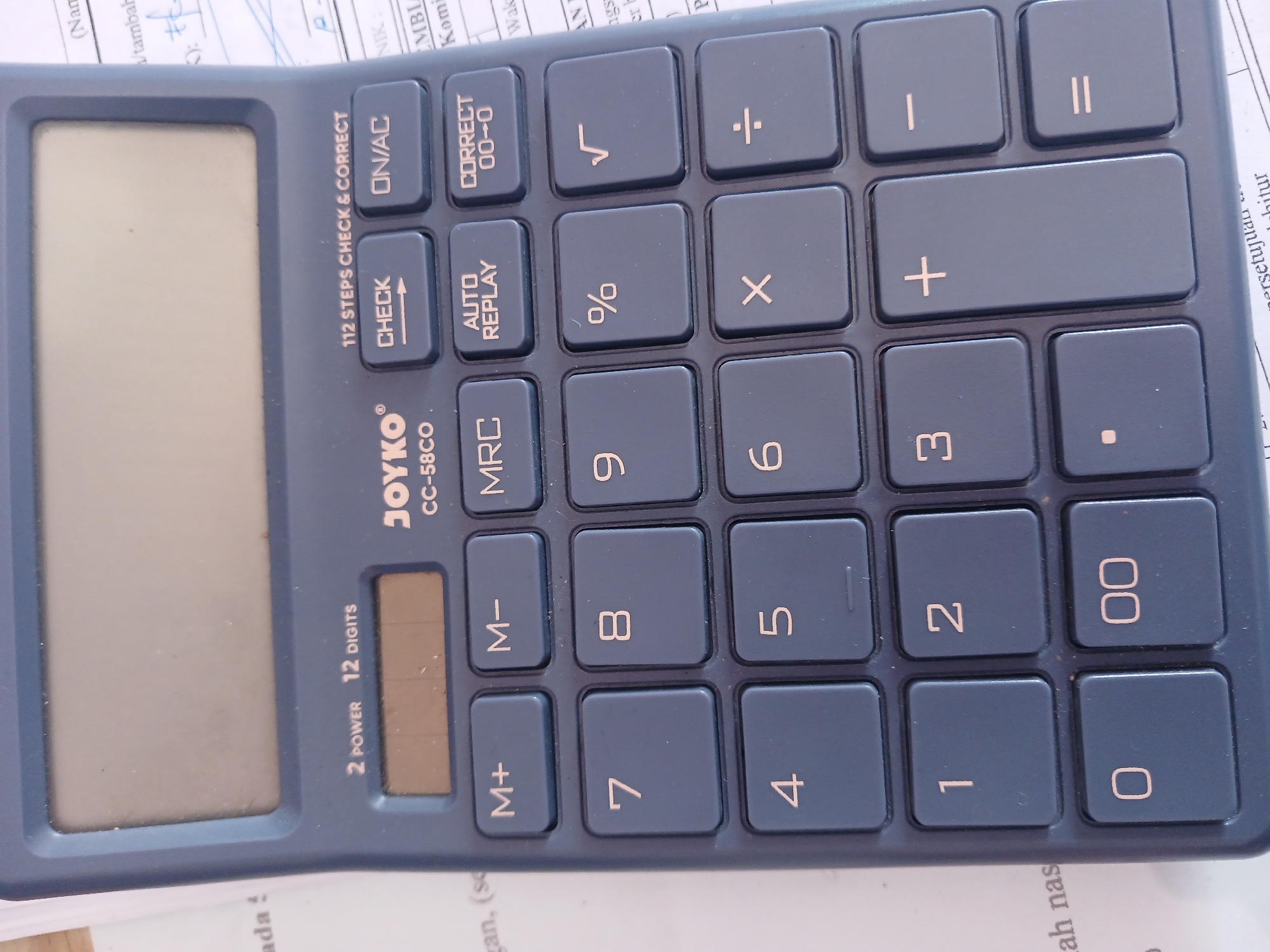 The image shows a close-up of a dark blue calculator, a Joyko CC-58CO model. The calculator is positioned on top of some papers, with some text visible on the papers, including the words "ada !" and "ah nas". The calculator has a large, blank digital display at the top. Below the display are several buttons, including "2 POWER 12 DIGITS", "CHECK", "ON/AC", "M+", "M-", "MRC", "AUTO REPLAY", and "CORRECT 00-0". The numerical keypad is also visible, with buttons for digits 0-9, ".", "00", "+", "-", "x", "÷", "=", and "%". The calculator appears to be in good condition, with no visible damage. The lighting in the image is consistent with indoor lighting. There is no identifiable information regarding the specific location within Watugede, Indonesia, as the image focuses solely on the calculator and its immediate surroundings.