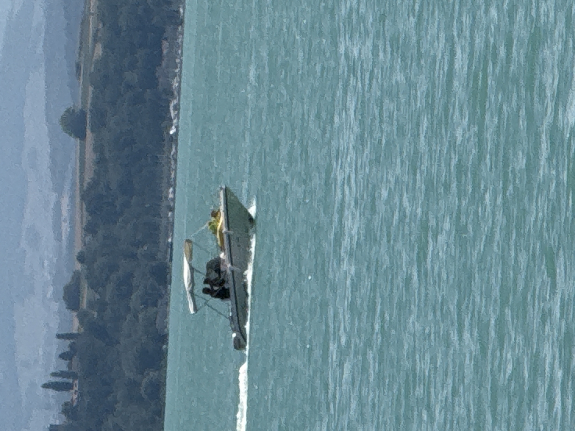 A motorboat with a canopy is moving across a body of water, leaving a wake. There are at least two individuals visible in the boat. In the background, a tree-covered hillside rises, with some buildings and structures visible on the crest of the hill. The sky is overcast and hazy, obscuring the distant landscape. The scene is consistent with a view of Lake Neuchâtel, Switzerland.
