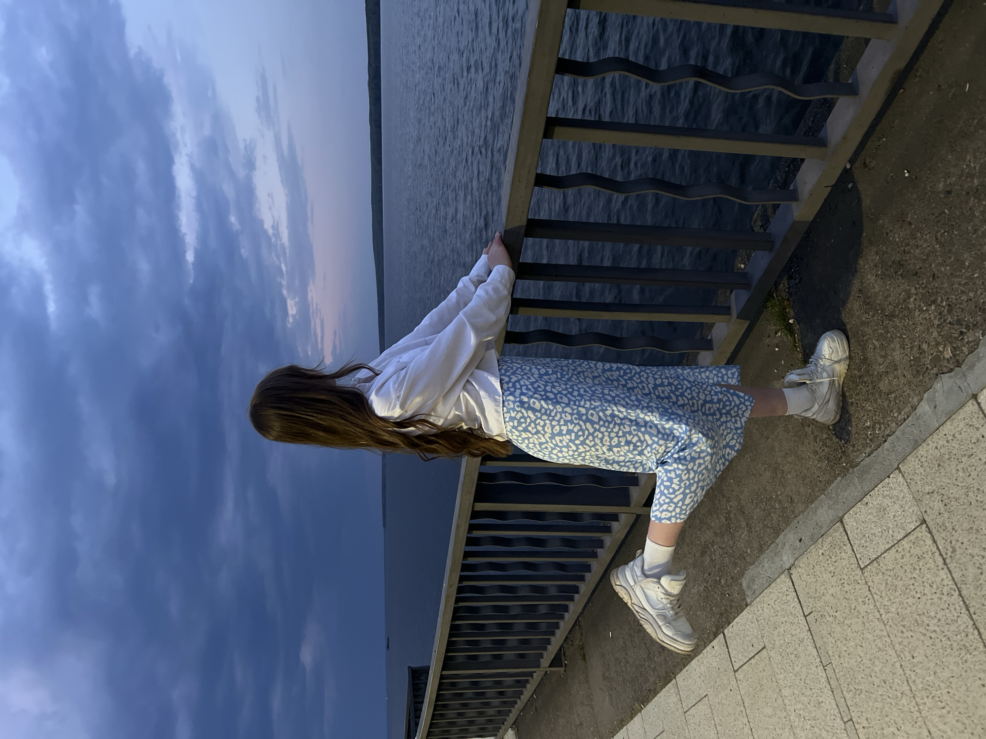 A young woman with long brown hair stands by a metal railing, overlooking a body of water under a dusk sky. She wears a white sweatshirt, a blue and white patterned skirt, white socks, and white sneakers. The railing is constructed of horizontal bars. The ground is paved with concrete. The water and sky fill the background, with the sky showing a gradient of blue to light pink near the horizon. The scene appears to be in the city of Chaikovsky, Russia.