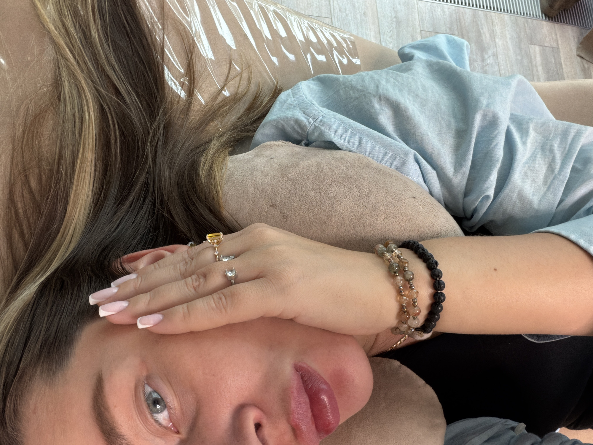 A woman lies on a beige, cushioned surface, her head resting on a matching pillow. She covers part of her face with her left hand. The woman's hair is a mix of dark and light brown. She wears a light blue button-down shirt and a black top. She has two rings on her left hand and two beaded bracelets on her left wrist. The setting appears to be indoors with a light-colored wooden floor visible in the background. A clear plastic sheet is partly visible near the top of the frame. The location is Unknown, Unknown.
