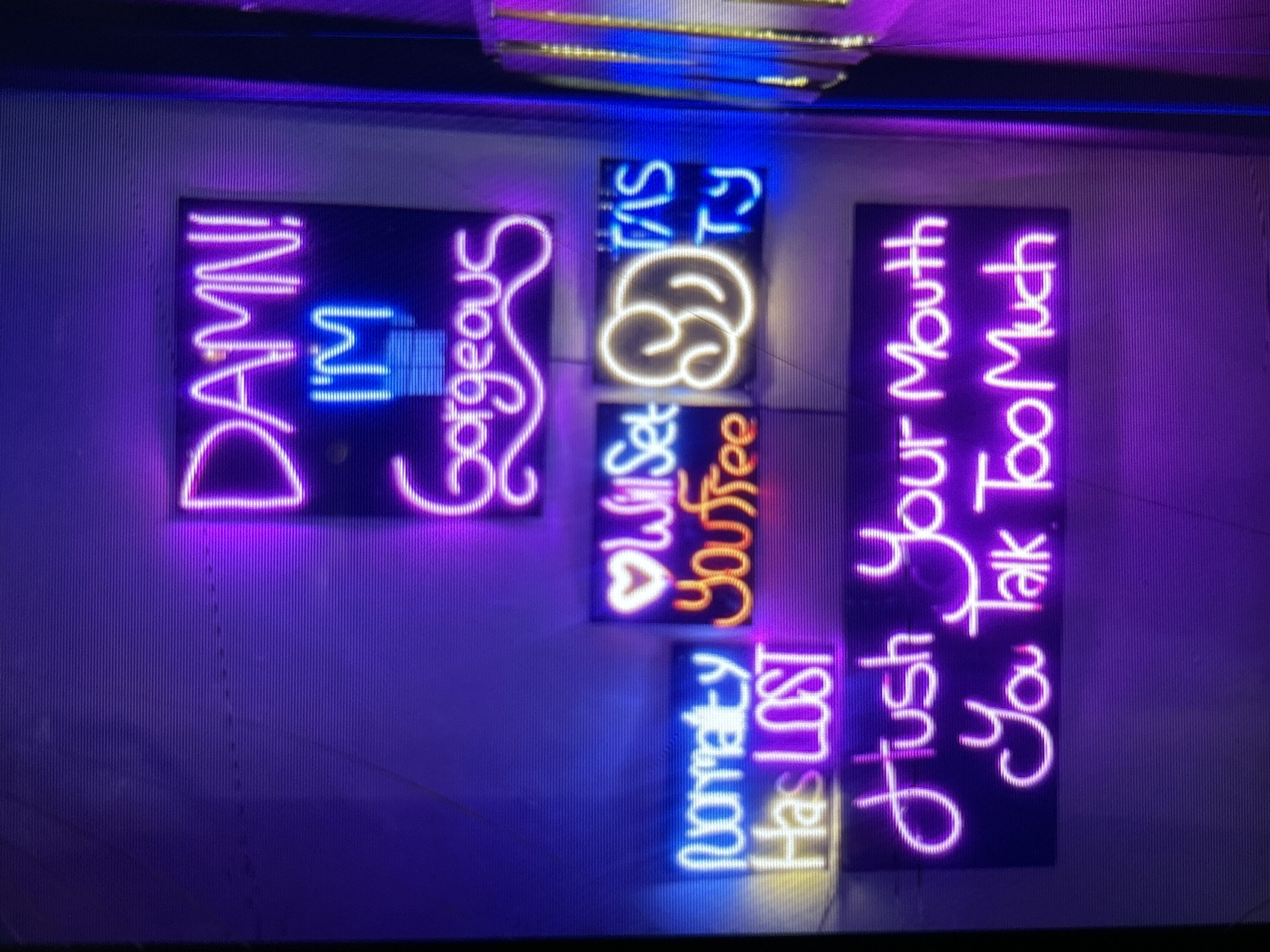 The image shows several neon signs with text. The largest sign at the top reads "DAMN! I'M GORGEOUS" in pink and blue neon. Below it, to the left, is a sign that says "Honesty Has LOST" in blue and pink neon. To the right of that, there is a small sign with a heart and the words "Will Set You Free" in pink and orange neon. Next to that, there is a sign with a yellow outline of what appears to be a swirling shape and the words "Tasty" in blue neon. The bottom sign reads "Hush Your Mouth You Talk Too Much" in pink neon. The signs are mounted on a purple wall. The overall lighting is dim, with the neon signs being the primary light source. The context of the image within Depok, Indonesia is not discernible from the visual information provided.