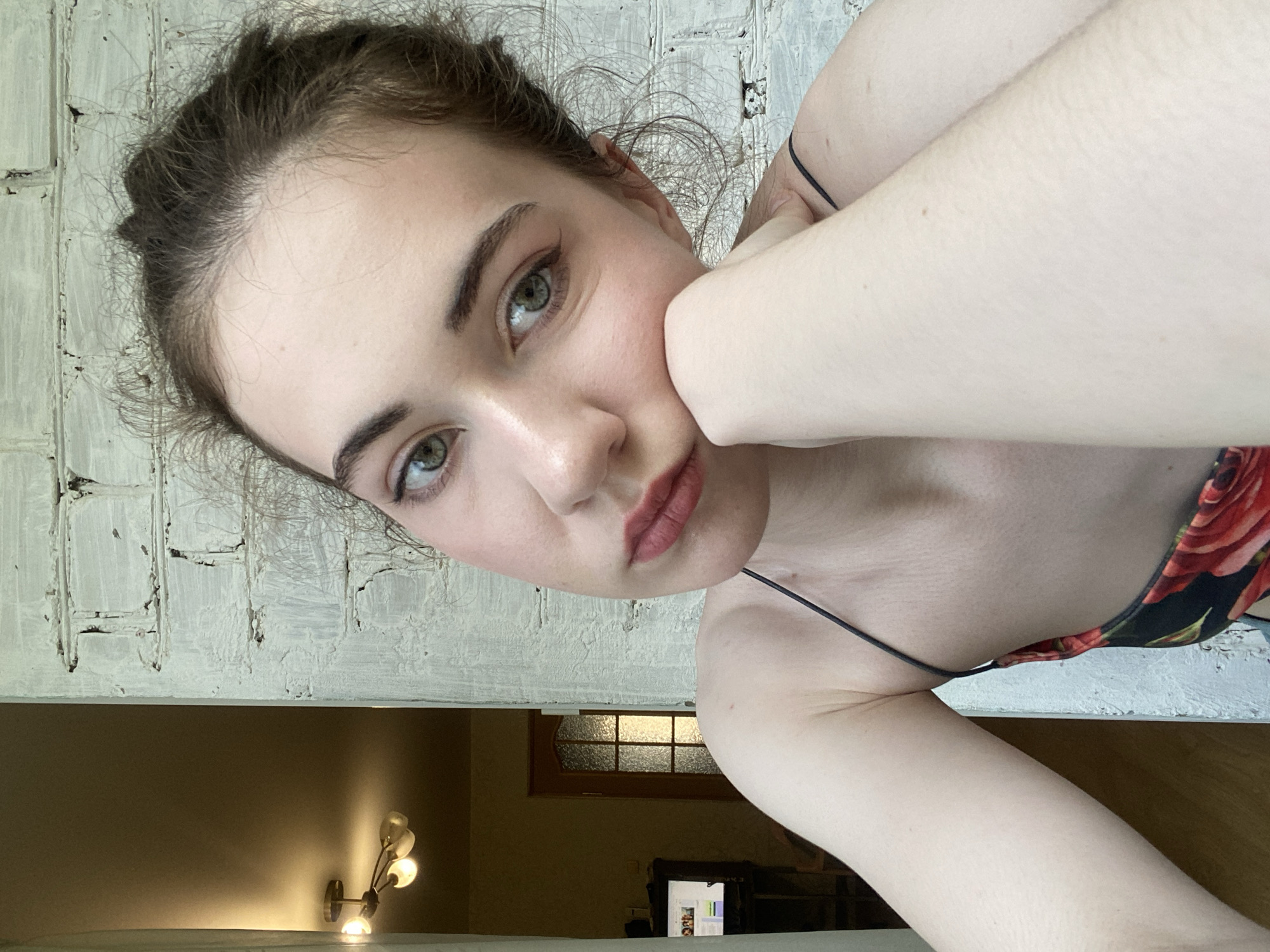 A young woman with light skin and brown hair tied up is shown in a close-up, selfie-style portrait. She is resting her chin on her right hand, with her left arm visible. She is wearing a black camisole with a red floral pattern. Her eyes are light green, and she is wearing light makeup, including lipstick. The background consists of a white brick wall and a doorway leading to an interior space with a chandelier visible. The image appears to have been taken indoors, near a window. The location is not Zavodoukovsk, Russia.