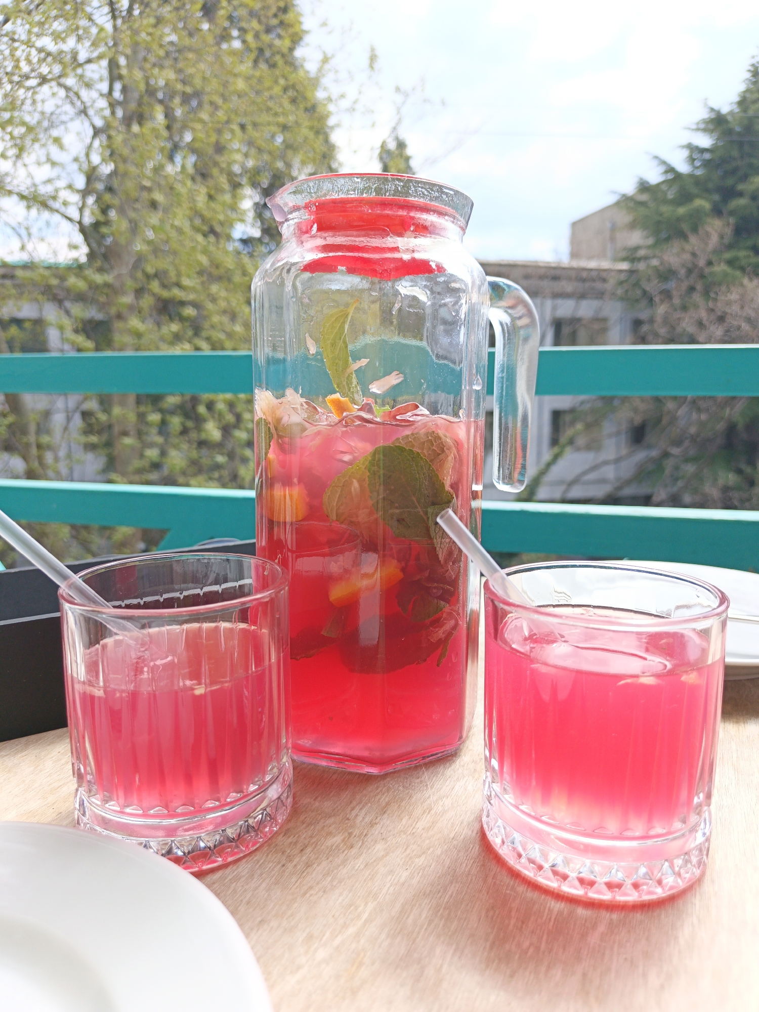 On a wooden table, a glass pitcher filled with a pink drink, adorned with mint leaves and orange slices, stands between two matching glasses also filled with the pink drink. A clear straw is placed in each container. The backdrop features a bright green fence with trees in the distance, suggesting an outdoor location. The scene is set in daylight, possibly on a balcony or patio, with Каштановское сельское поселение, Ukraine serving as the potential city context. A partial view of a white plate can be seen on the table.