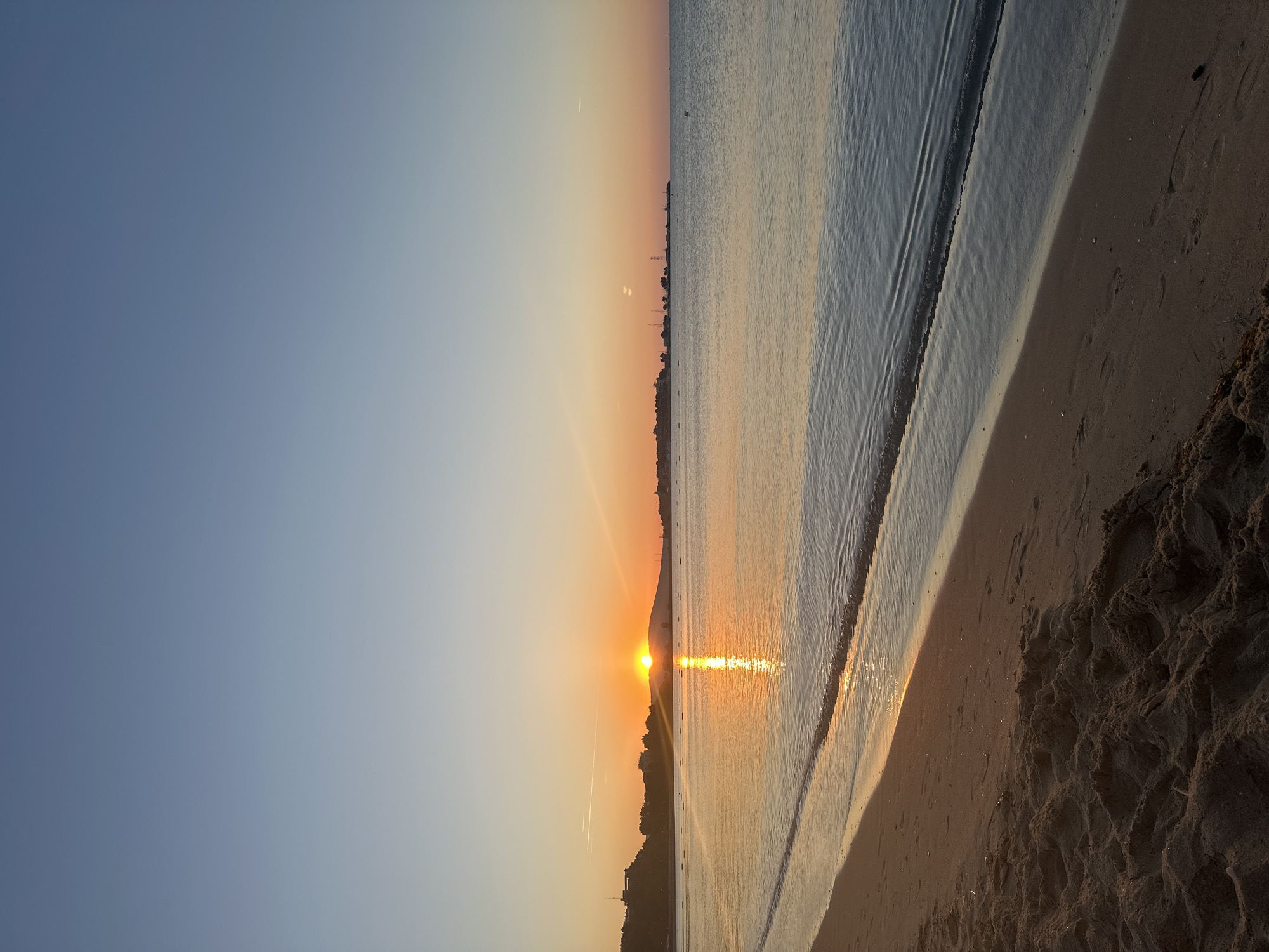 The image depicts a tranquil sunrise over the Black Sea, viewed from the sandy shore of Nesebar, Bulgaria. The sky transitions from a deep blue at the top to a soft orange and yellow hue near the horizon, where the sun is just beginning to rise behind a distant, silhouetted hill. The sun's rays cast a shimmering golden path across the calm water, reflecting the warm colors of the dawn.
The foreground is dominated by the sandy beach, with subtle textures and footprints visible in the sand. The gentle waves of the sea lap at the shoreline, creating a delicate ripple effect. In the mid-ground, a low-lying coastline is visible, dotted with buildings and structures, hinting at the historic town of Nesebar. A few small boats or buoys are scattered across the water. The overall atmosphere is serene and peaceful, characteristic of an early morning on the Bulgarian coast.