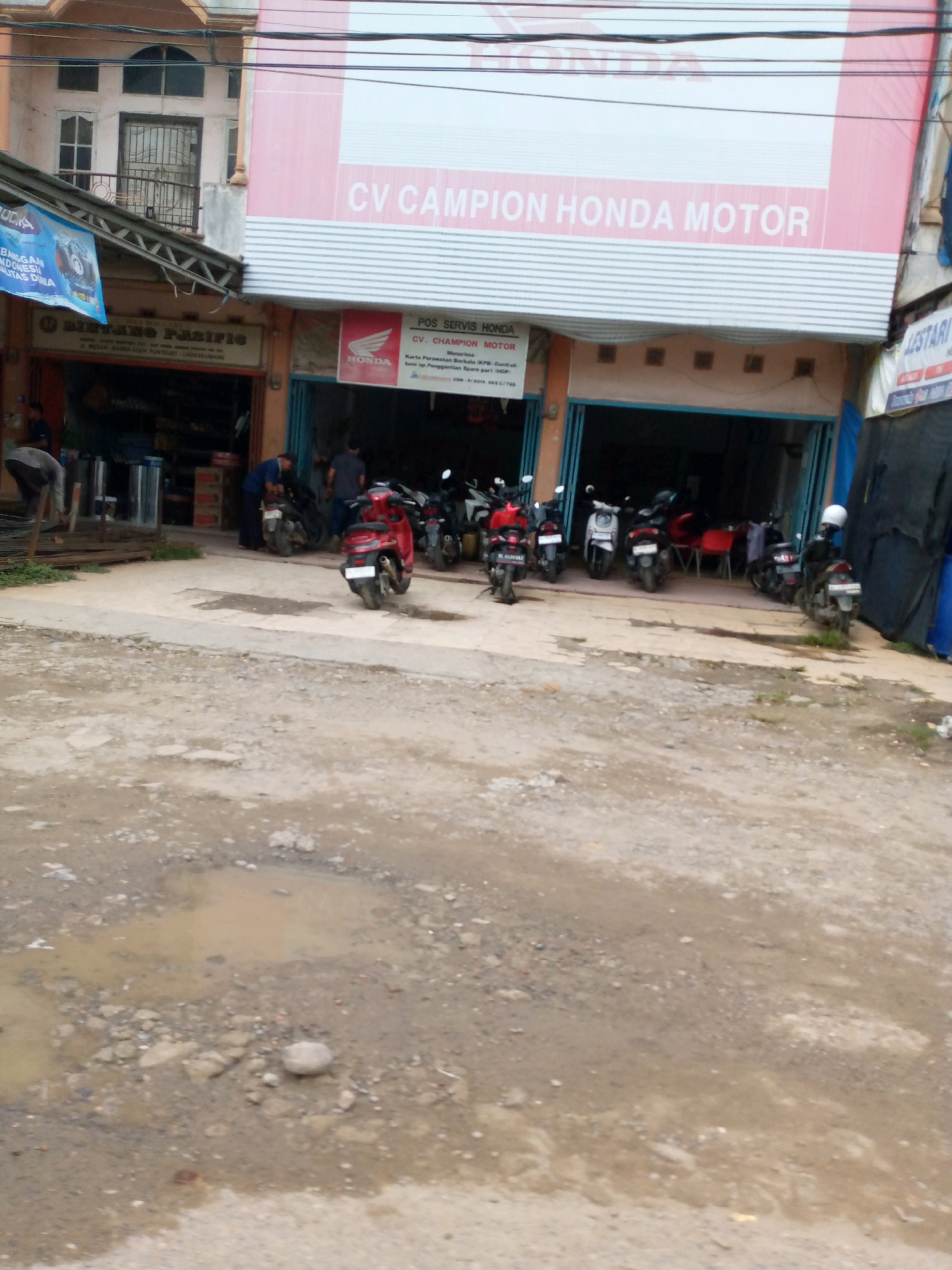This is a medium-distance, eye-level, full-shot photograph of the exterior of a Honda motorcycle dealership in Lhokseumawe, Indonesia. The dealership, identified by a large pink sign reading "CV CAMPION HONDA MOTOR," is situated on a street with a potholed and muddy asphalt surface in the foreground. The building has an open-front design with a covered walkway where several Honda motorcycles are parked, both inside the open bays and on the sidewalk. Two individuals are visible inside the dealership, one appearing to be working on a motorcycle, and another person is standing near the entrance. A third person is seen from behind, leaning on a piece of metal construction material on the left side of the frame. A blue banner with white lettering is hanging on the left side of the building. The environment appears to be a commercial street with other businesses visible.