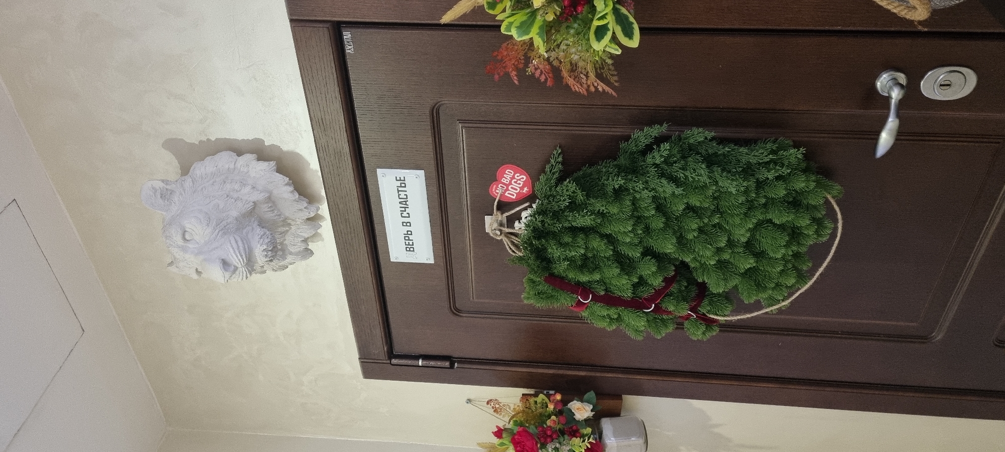 The image shows a dark brown wooden door with a wreath of green fir branches hanging on it. A small, heart-shaped red tag with the text "NO BAD DOGS" is attached to the wreath. Below the wreath, a white rectangular sign with the Russian text "ДВЕРЬ В СЧАСТЬЕ" (Door to Happiness) is affixed to the door. To the left of the door, on a textured beige wall, is a white decorative plaster relief of a wild boar's head. A metallic door handle and a keyhole are visible on the door to the right of the wreath. At the bottom of the image, a small arrangement of artificial flowers and berries is visible, suggesting a festive or decorative context. The lighting indicates it is likely indoors, and the overall impression is one of home decoration, possibly for a holiday or as a permanent fixture. The location is specified as Moscow, Russia.