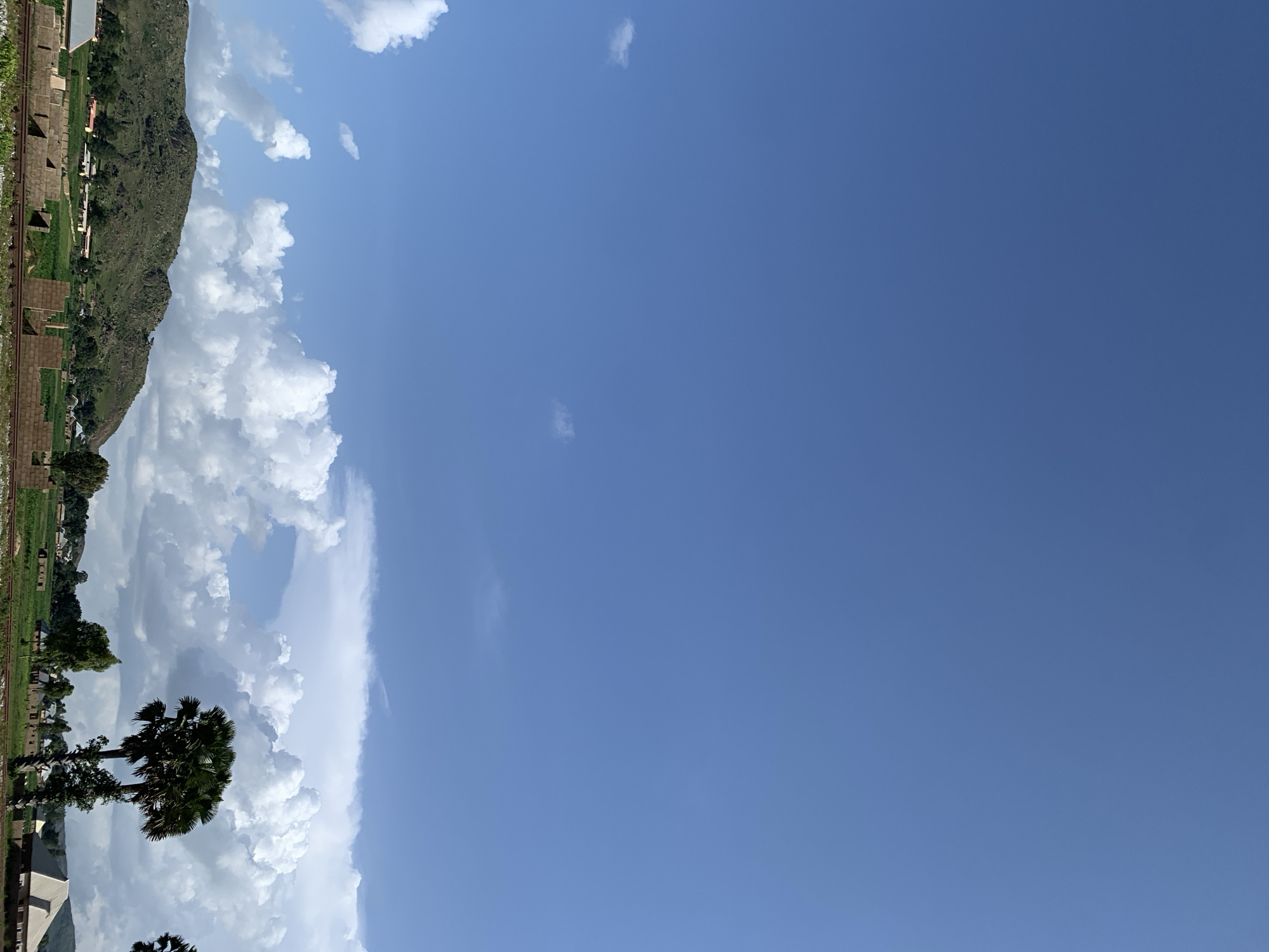 The image depicts a rural landscape in Kuru Karama, Nigeria, under a vast blue sky with scattered white clouds. In the foreground, unfinished brick structures are visible, suggesting ongoing construction. A railway track runs horizontally across the bottom of the frame. Beyond the construction, a green field stretches out, dotted with small buildings and trees. A prominent hill, covered in vegetation and rocks, rises in the mid-ground. Two palm trees stand out on the right side of the image, with lush green foliage. The overall scene is tranquil and indicative of a developing rural community.