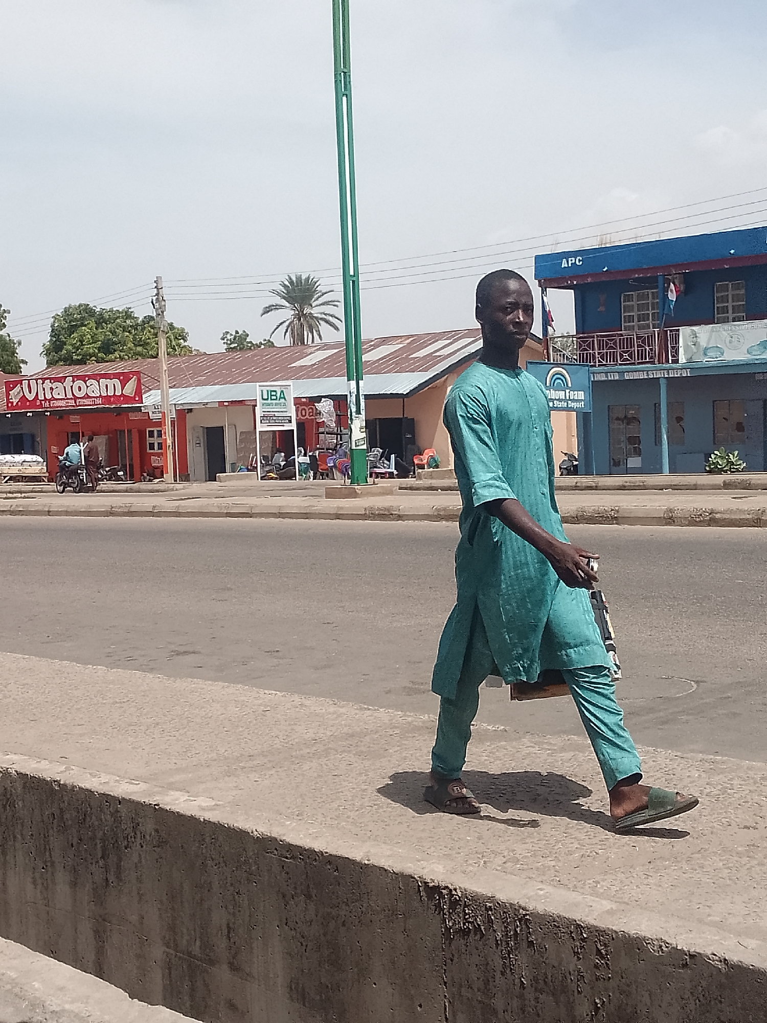 A medium shot shows a man walking on a curb next to a street. He is wearing a turquoise outfit, consisting of a long-sleeved tunic and pants, and slip-on sandals. He holds a dark-colored object in his right hand. The street is flanked by buildings. On the left, there is a red building with a sign that says "Ultafoam" and another sign that reads "UBA." People are gathered outside. In the background, a blue building is visible with the letters "APC" on top. The words "Rainbow Foam LTD. Gombe State Depot" are on the building. A green metal pole stands next to the curb. The image appears to be taken in Pantame, Nigeria.
