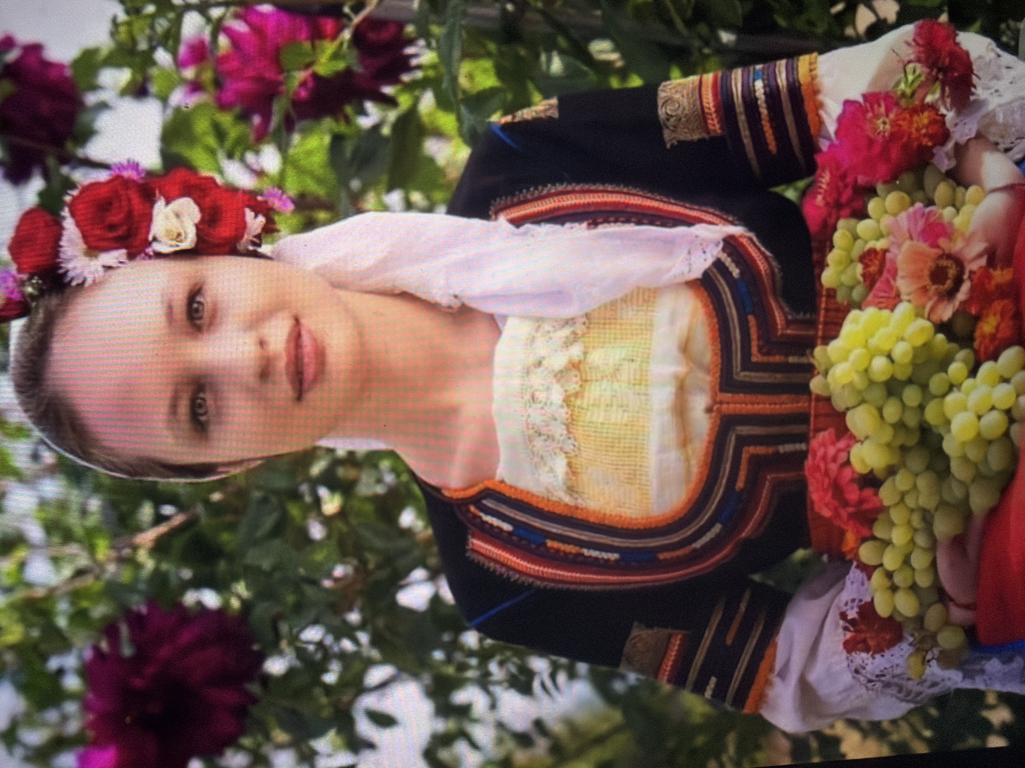 A young adult female is depicted in a chest-up portrait, gazing directly at the viewer with a neutral expression. Her hair is drawn back, and she wears a white headscarf with a cluster of red roses and smaller white and pink flowers positioned on the left side of her head. Her attire consists of a long-sleeved white blouse with lace cuffs, an undergarment featuring a cream-colored, textured bodice, and a dark-colored jacket or vest. This outer garment is adorned with intricate multi-colored embroidery and trim (red, orange, blue, gold, black) arranged in geometric patterns around the neckline and on the sleeves. She holds a shallow, red container at chest level, filled with several bunches of light green grapes and various cut flowers, including red, pink, and orange blooms. The background, blurred, features dense green foliage and large, dark pink/purple flowers, suggesting an outdoor, vegetated location. This scene takes place in the city of Shumen, Bulgaria.