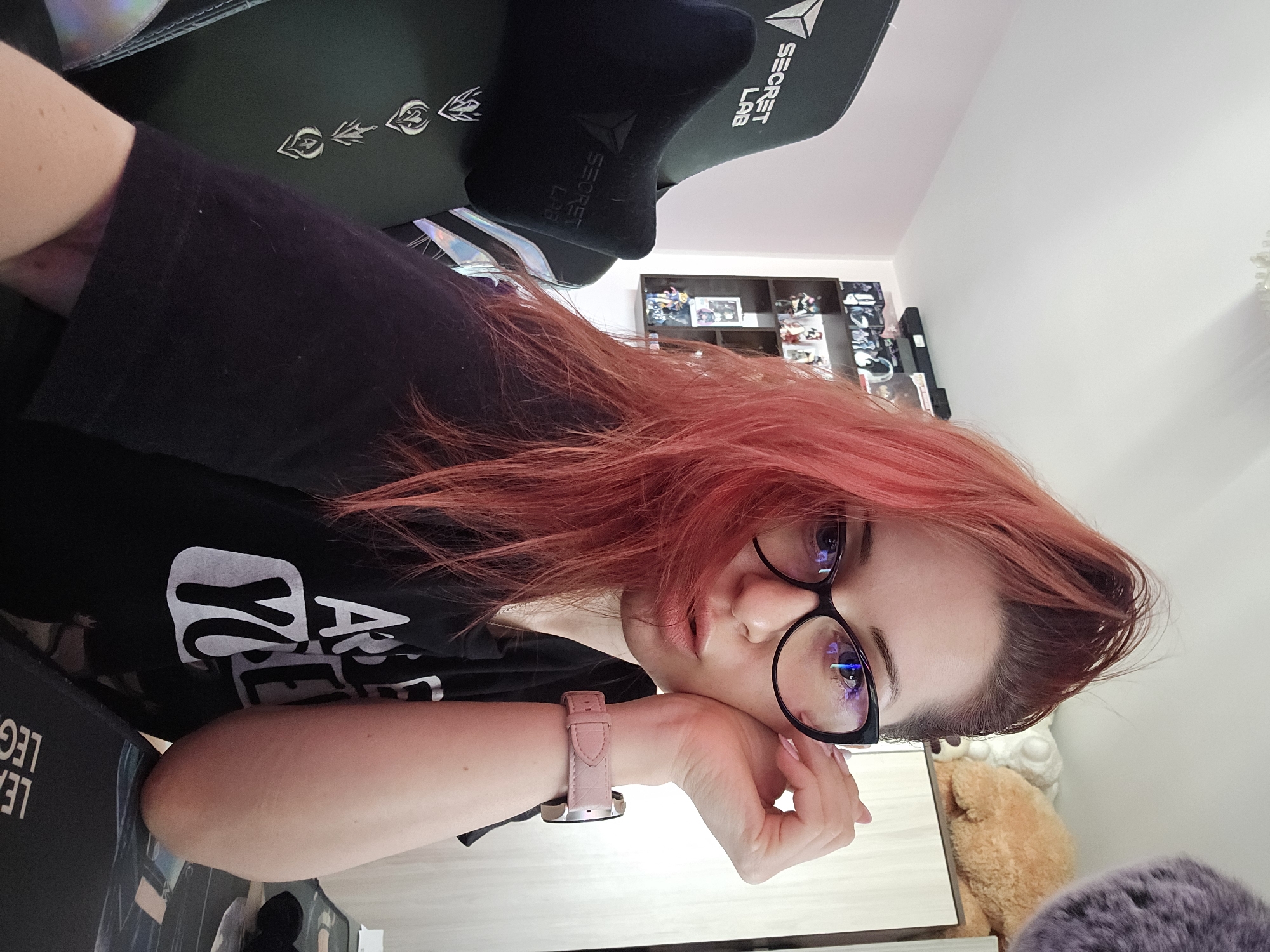 The image depicts a medium shot of a young woman with vibrant red hair, wearing black-rimmed glasses and a black t-shirt with white text. She is seated in a dark green gaming chair with "SECRET LAB" branding and a black headrest. Her right hand is resting on her cheek, and she is wearing a pink watch on her left wrist. In the background, there is a shelf filled with figurines and several stuffed animals, including a light brown bear. To the right, a closet or cabinet is visible. The overall setting appears to be a room in a residence, possibly a home office or gaming setup. The scene does not contain any discernible elements that would allow for precise identification of a location within the city of Burgas, Bulgaria.