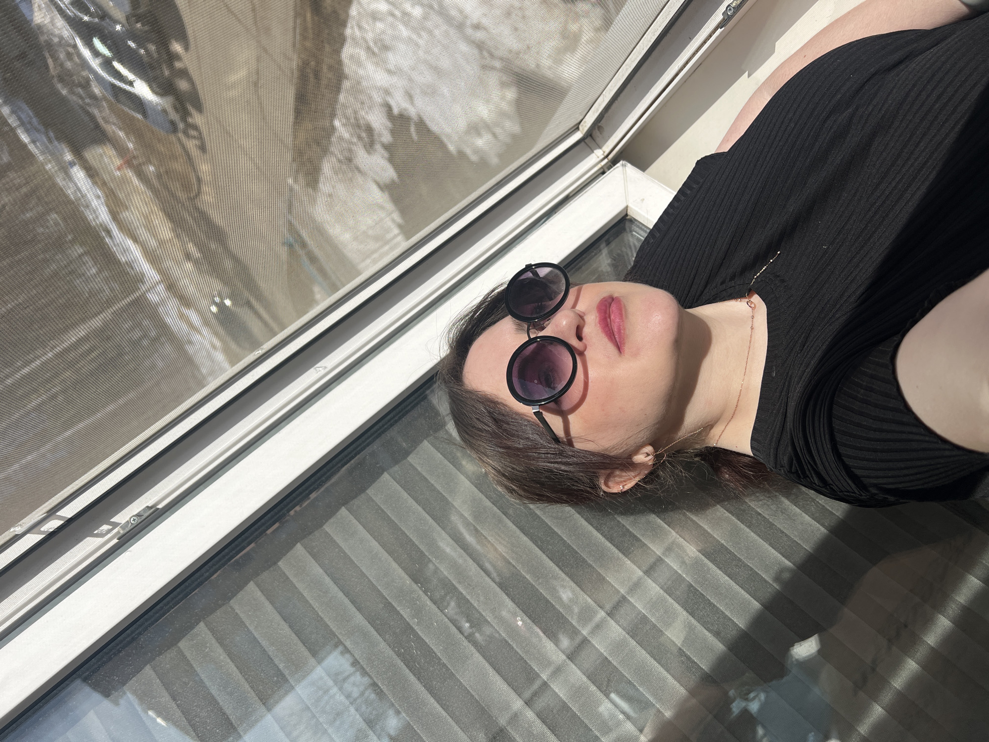 A woman wearing sunglasses and a black shirt is lying near a window. The window has a screen and blinds. The location is not identifiable as Noviy, Russia based on the visual information provided.