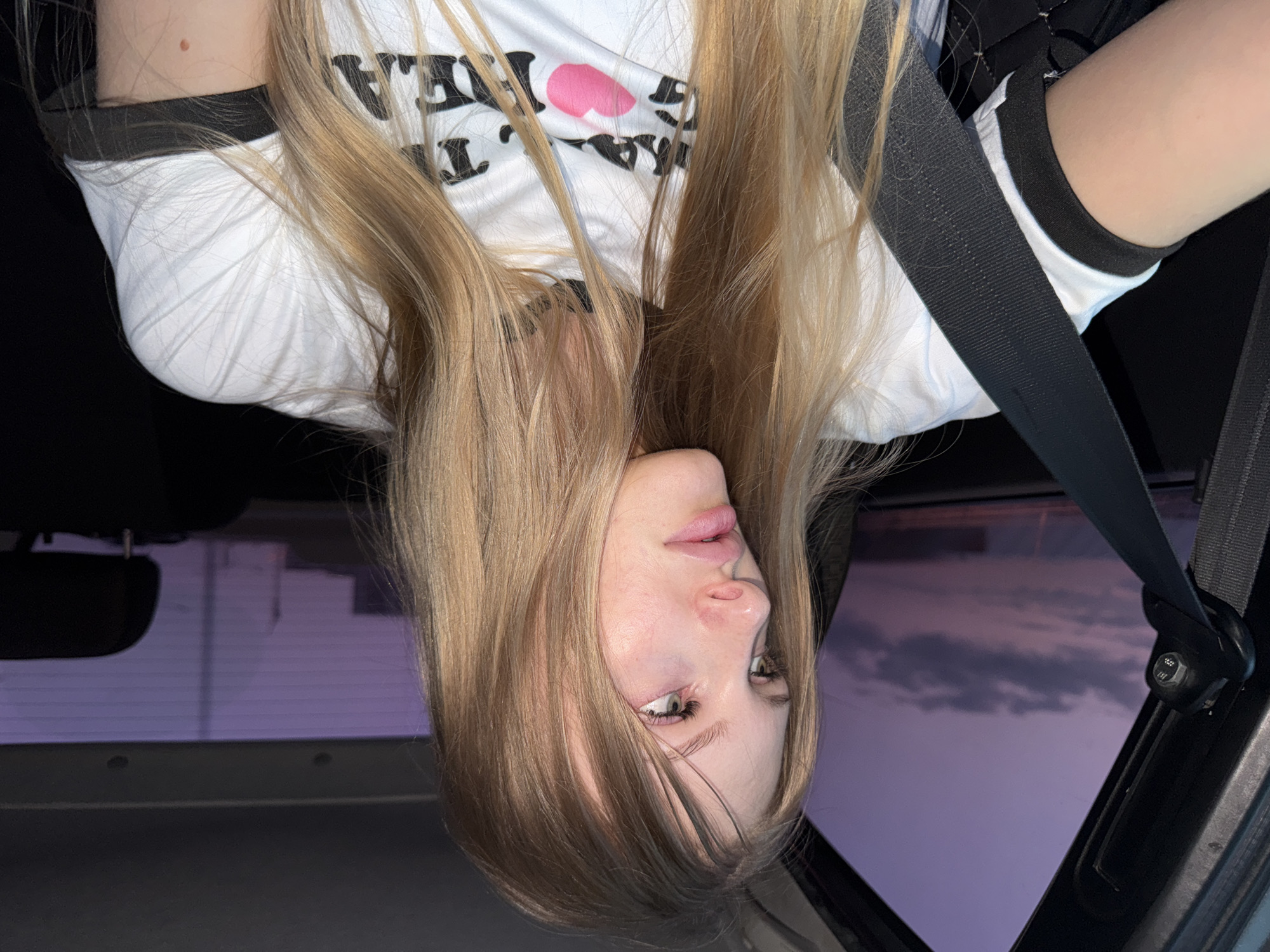 A young woman with long blonde hair is seen from the chest up inside a car, looking out the window. The sky outside is a deep purple, indicating either dawn or dusk, with some scattered clouds. She is wearing a white t-shirt with a black collar and a graphic print on the front that includes the text "MAKE. THE. HEART." above a pink heart graphic. A black seatbelt crosses over her chest. The interior of the car is dark, with the headrest of a seat visible behind her. The scene appears to be set in the city of Perm, Russia, based on the provided context.