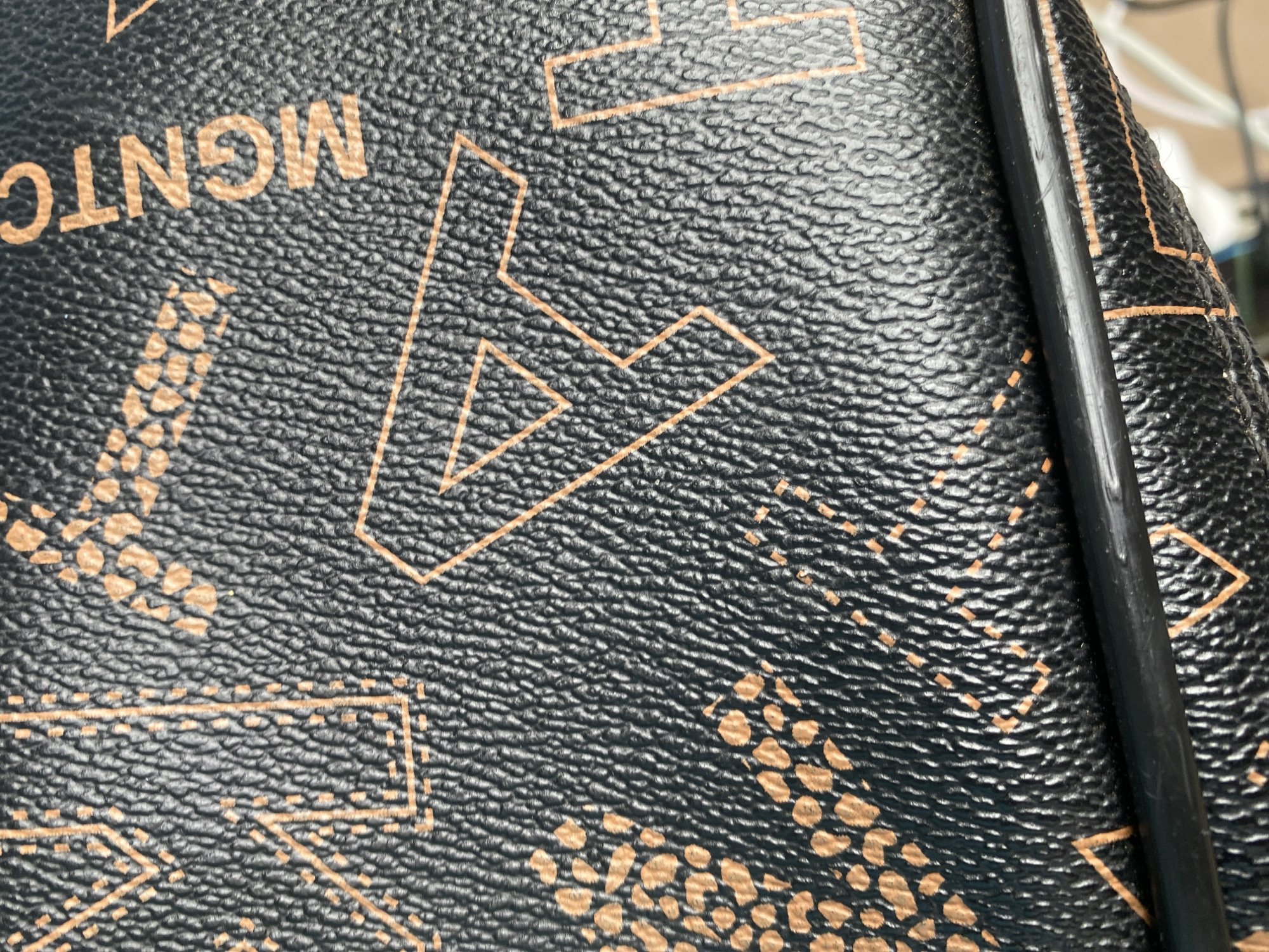 A close-up, overhead shot shows a black leather bag with an orange MCM logo pattern. The pattern features the letters "MCM" and diamond shapes. A thin black strap runs horizontally across the lower portion of the image. The textured surface of the leather is visible. The image appears to be taken indoors, with a blurred background suggesting other objects. No specific location in Jalingo, Nigeria is identifiable from the image.