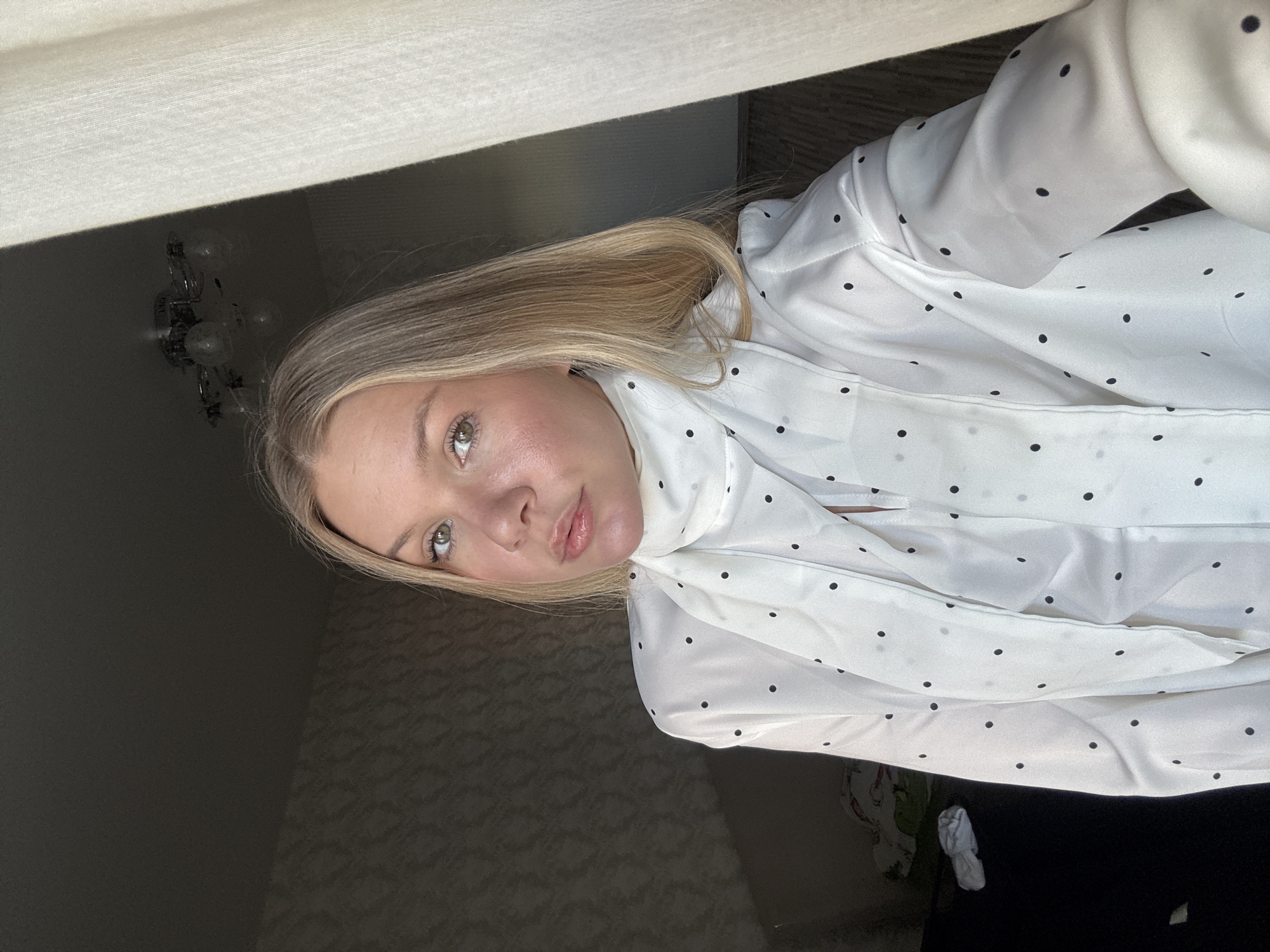 A woman with blonde hair is wearing a white blouse with black polka dots. She is indoors. A chandelier is visible on the ceiling in the background. The location is most likely in Perm, Russia.