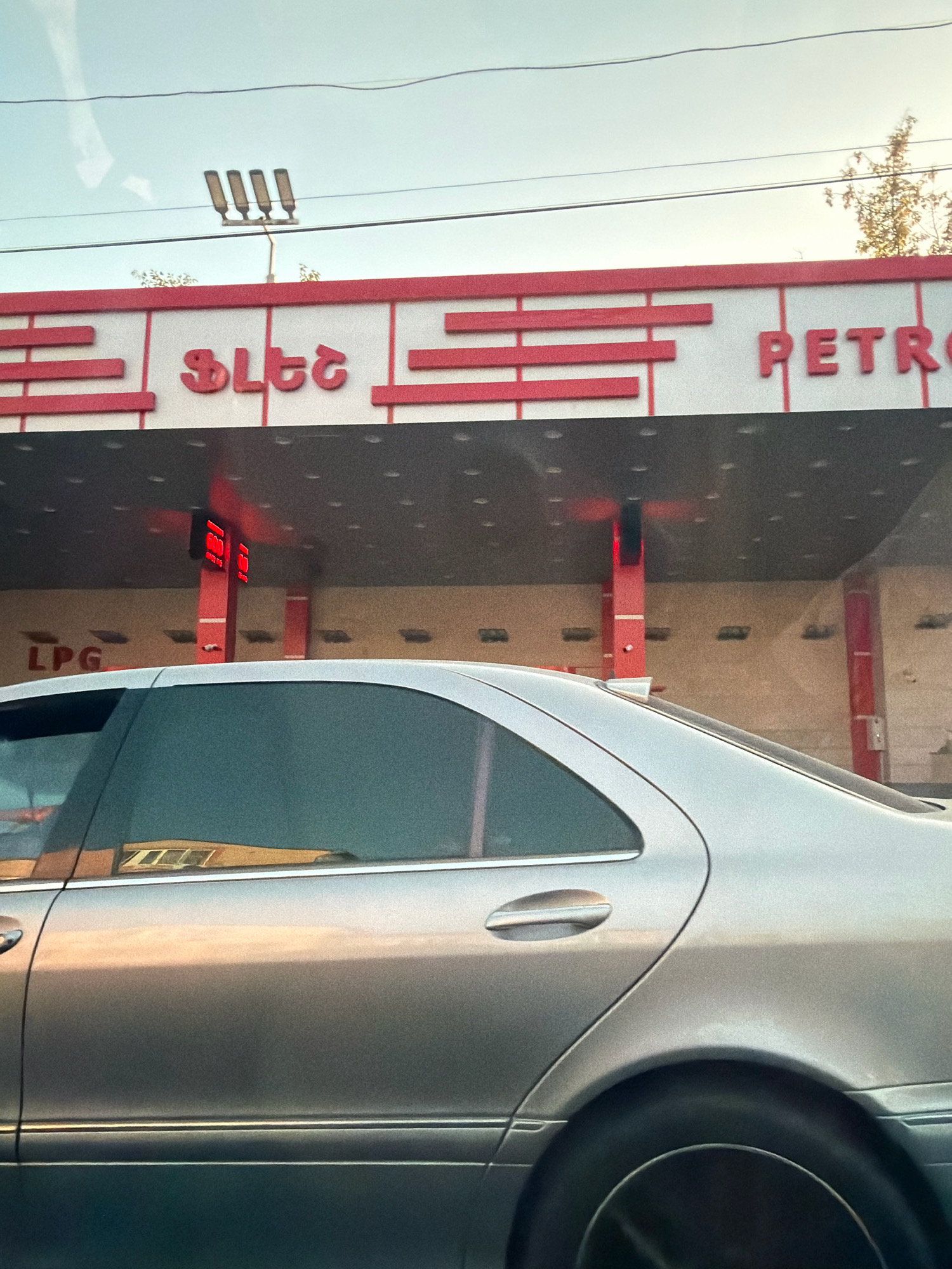 A silver sedan is parked at a gas station under an overhang. The gas station has a red and white facade with a red sign reading "PETRO". There are red rectangular lights indicating prices for gas. A sign for "LPG" is visible on the left side of the overhang. The sky is clear and a few trees are visible in the background. The image appears to have been taken during the day.