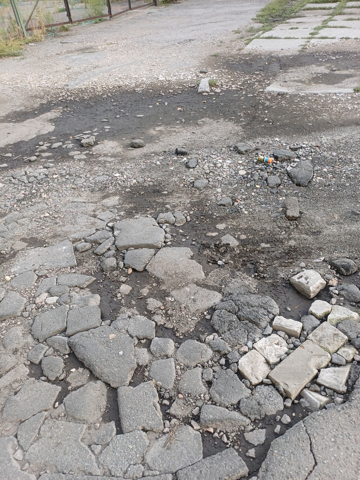 The image depicts a heavily damaged asphalt road with numerous potholes and broken sections. The road surface is a mix of asphalt, gravel, and dirt, with some areas appearing to have standing water or mud. There are also several pieces of concrete debris scattered across the road. In the background, a metal fence and some vegetation are visible, suggesting an outdoor location. A few small colorful objects, possibly litter such as plastic caps or wrappers, are visible amongst the debris. The overall scene conveys a sense of disrepair and neglect. The image does not show any people, animals, or specific landmarks that would allow for a precise location within Kyiv, Ukraine.