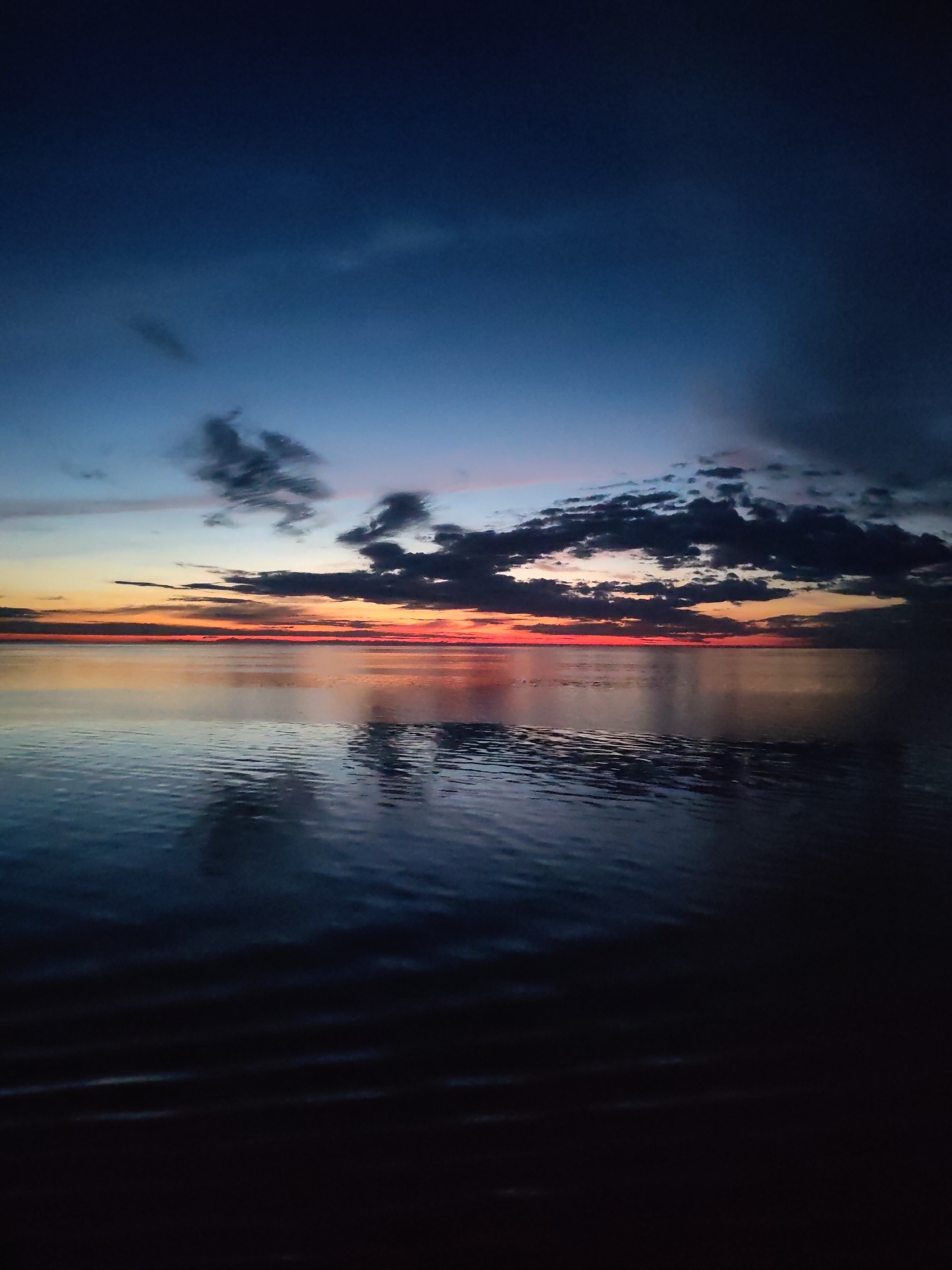The image depicts a body of water, likely a lake or river, under a twilight sky. The sky transitions from deep blue to a mix of orange and red near the horizon, where the sun has recently set. Dark, puffy clouds are scattered across the upper portion of the sky and clustered along the horizon. The water reflects the colors of the sky and clouds, creating a mirrored effect, with gentle ripples disturbing the reflection. The foreground is obscured in shadow. No people or identifiable objects are visible in the scene. There is no information to confirm that this photo was taken at the city of Mologa, Russia.