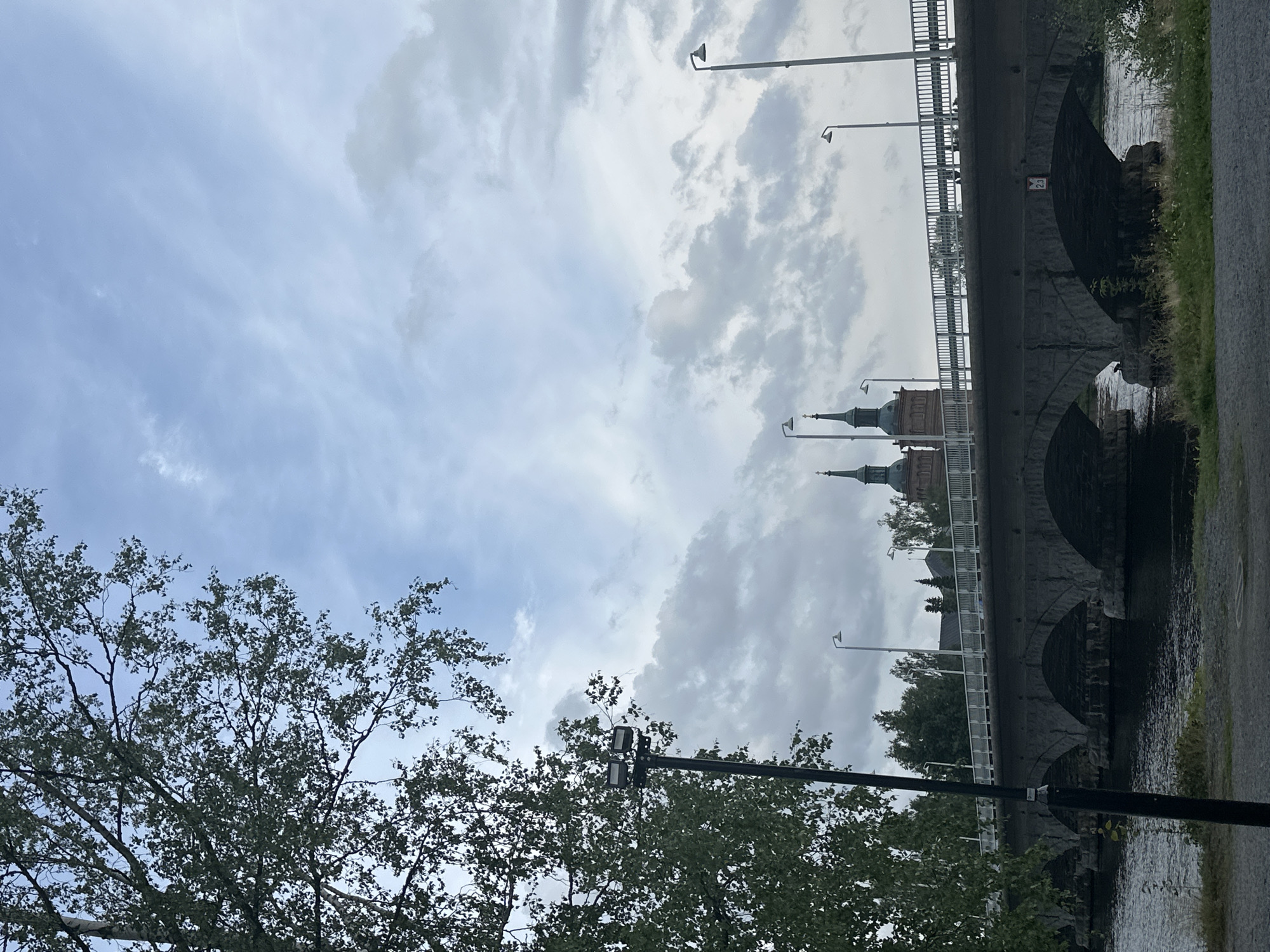 The image captures a view of the bridge and river in Vammala, Finland. A large, multi-arched stone bridge spans a dark, flowing river in the mid-ground. The bridge has a white railing along its top, and streetlights are visible along its length. On the far side of the river, behind the bridge, are trees and two prominent church spires, suggesting a historic or religious center. The sky is cloudy with patches of lighter blue and darker gray clouds. In the foreground, to the left, a large tree with green leaves partially obscures the view. A dark lamppost with two lights stands in the far left foreground. The overall scene suggests an overcast day in a European city with a body of water and significant architecture.