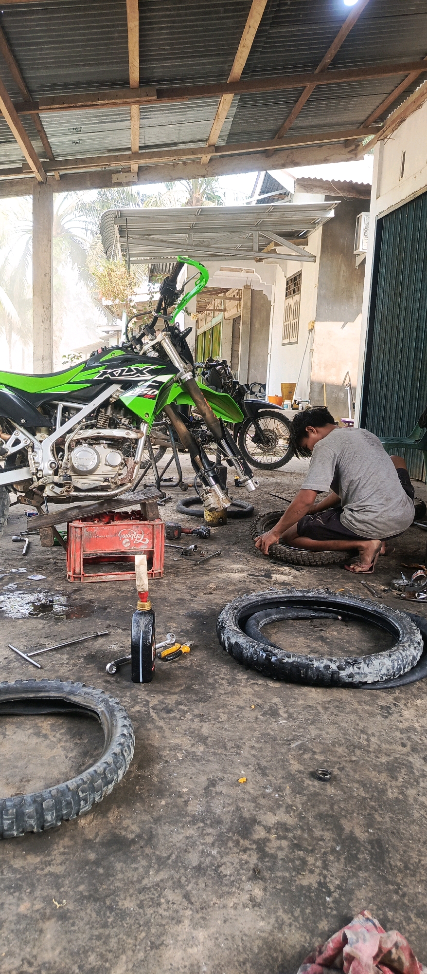 In Bangun Purba Barat, Indonesia, a man is working on a motorcycle tire in a workshop. The scene is outdoors under a corrugated metal roof supported by wooden beams. A green and black dirt bike, labeled "KLX 150," is prominently displayed, partially disassembled, with its front wheel removed. Another motorcycle, mostly black, is visible behind it. The ground is concrete, stained with oil and dirt. Tools, including wrenches and a tire lever, are scattered around. A red plastic crate sits near the dirt bike. A bottle of oil with a dispenser cap and a small yellow tool are on the ground. Two detached motorcycle tires are also present. The man, wearing a grey t-shirt and dark shorts, is kneeling and appears to be manipulating a tire, possibly changing or repairing it. His posture suggests focus on the task at hand. The background shows a partially open green garage door and a wall with a window. The overall impression is of an active, albeit somewhat cluttered, motorcycle repair environment.