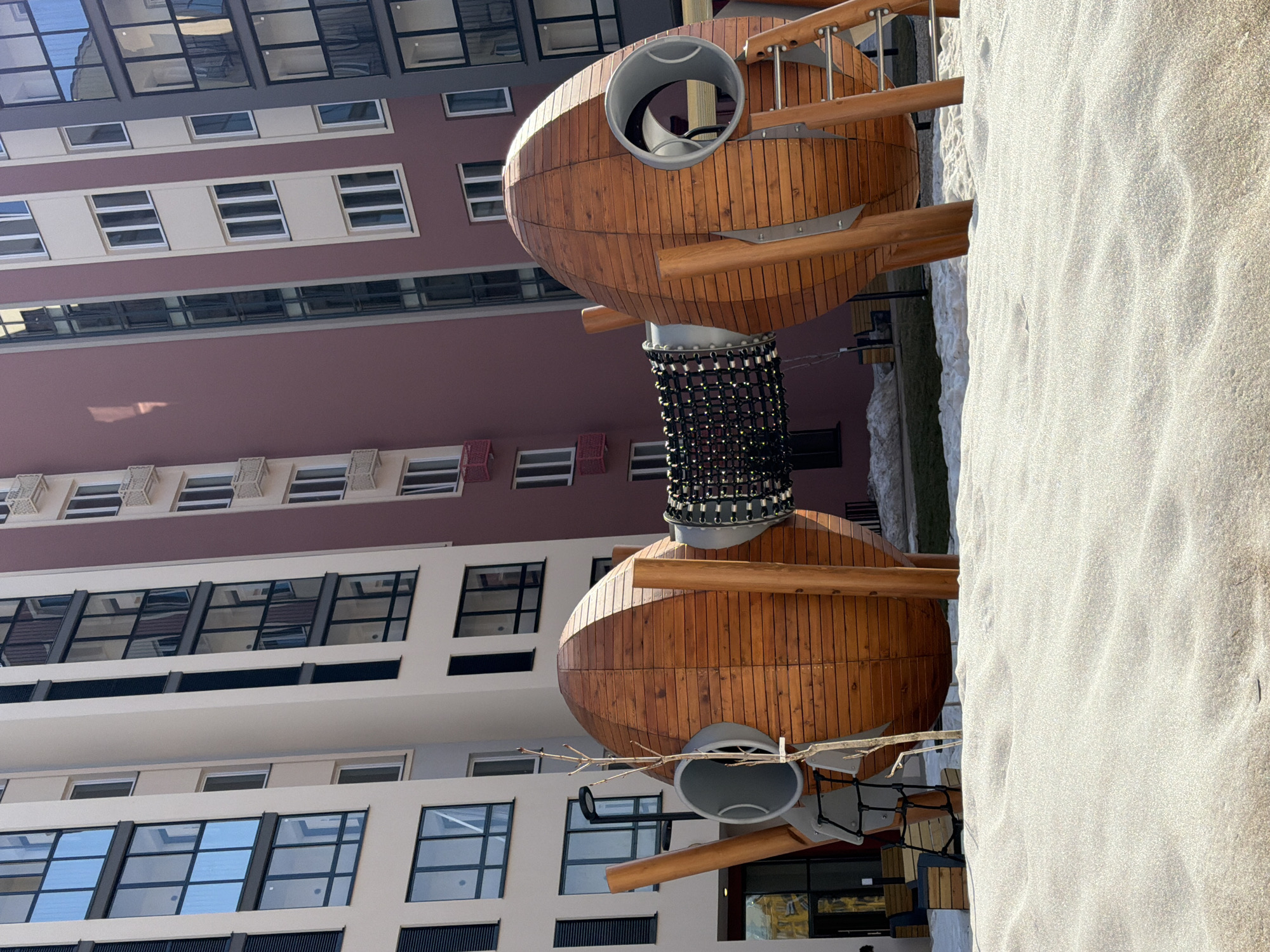 The image shows a wooden playground structure with two rounded, oval-shaped main sections connected by a cylindrical, net-covered tube. Each oval section has a metal opening on its side. The structure appears to be located in a sandbox area with light-colored sand. In the background, there is a multi-story building with a mix of architectural styles, featuring repeating window patterns and different colored sections. The location is Pervomaysky, Russia. There are no visible people or interactions in the scene. The environment appears to be a modern urban setting with a focus on recreational space.