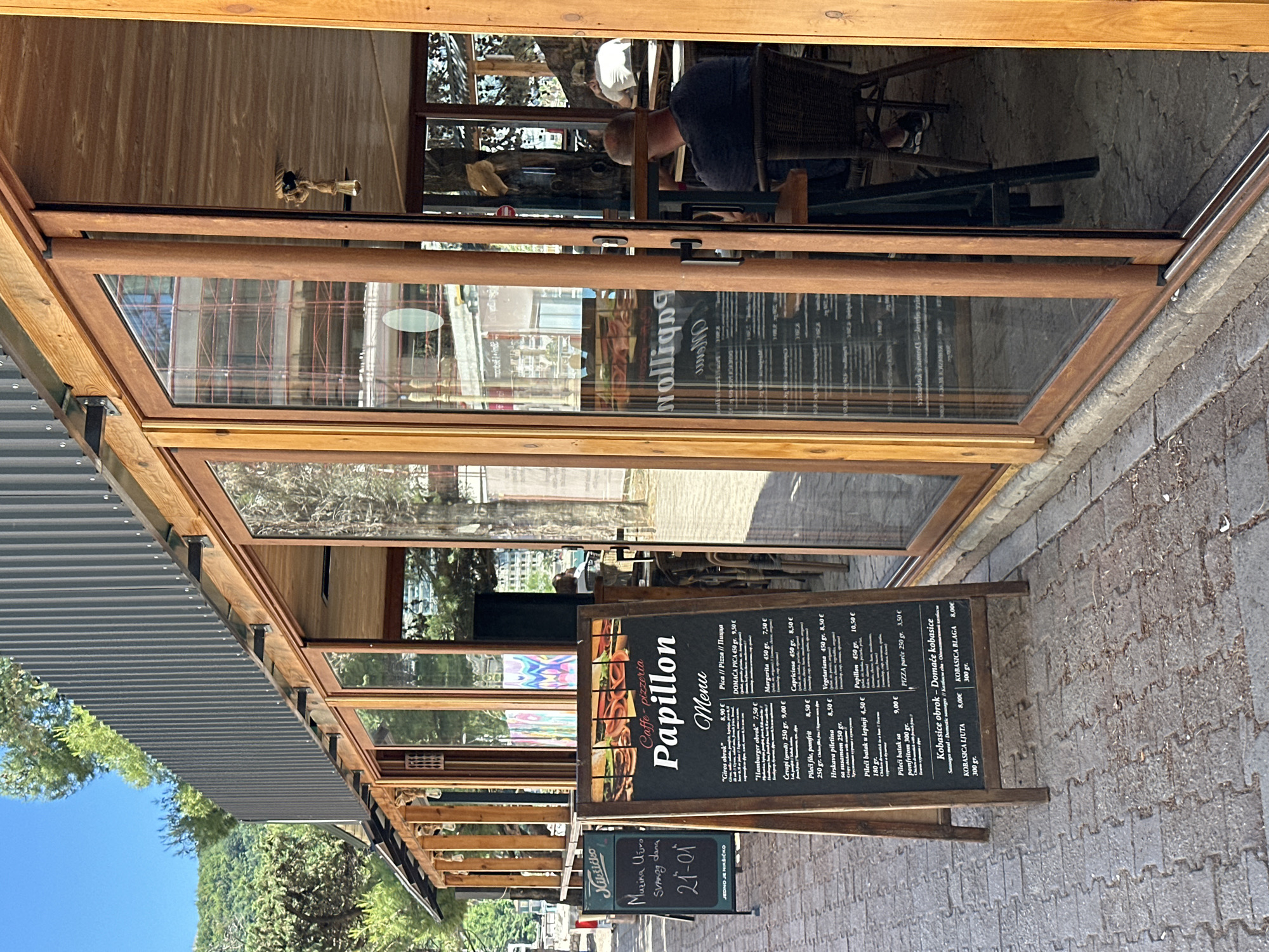 The image depicts the exterior of a restaurant named "Papillon" in Rafailovići, Montenegro. The restaurant has a partially covered seating area with a retractable roof and large folding glass doors. A chalkboard menu displays prices in Euros for various items including pizzas, "Giros obrok" (gyro plate), "Hamburger obrok" (hamburger plate), and other dishes. Another chalkboard displays "Nikšičko" as well as "Masina Vino Svakog dana", with "21-01" below. A man is seated at a table inside the restaurant, visible through the glass doors. The outdoor area is paved with gray interlocking stones. Trees and sky are visible in the background.