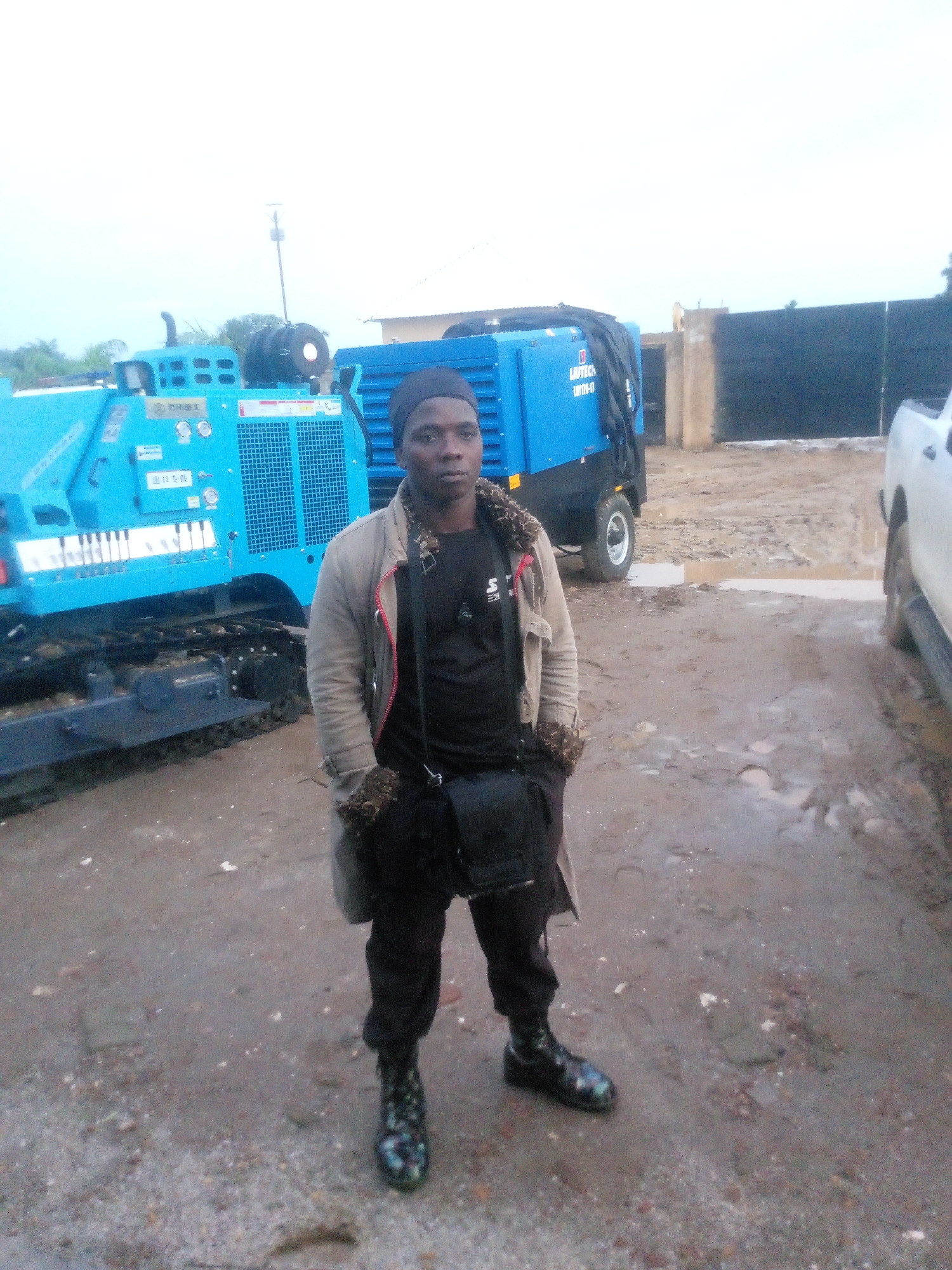 A medium shot shows a Black male standing in a muddy outdoor area. He is wearing a black beanie, a tan jacket with faux fur trim, a black t-shirt, and black pants. He has a black crossbody bag and is wearing black boots with a colorful, iridescent pattern. He is standing in front of a large blue drilling machine. To his right, there is a blue generator and a white pickup truck. The ground is covered in mud and puddles, suggesting recent rainfall. The background shows a fence and some vegetation. The sky is overcast. The location is identified as Indo, Nigeria.