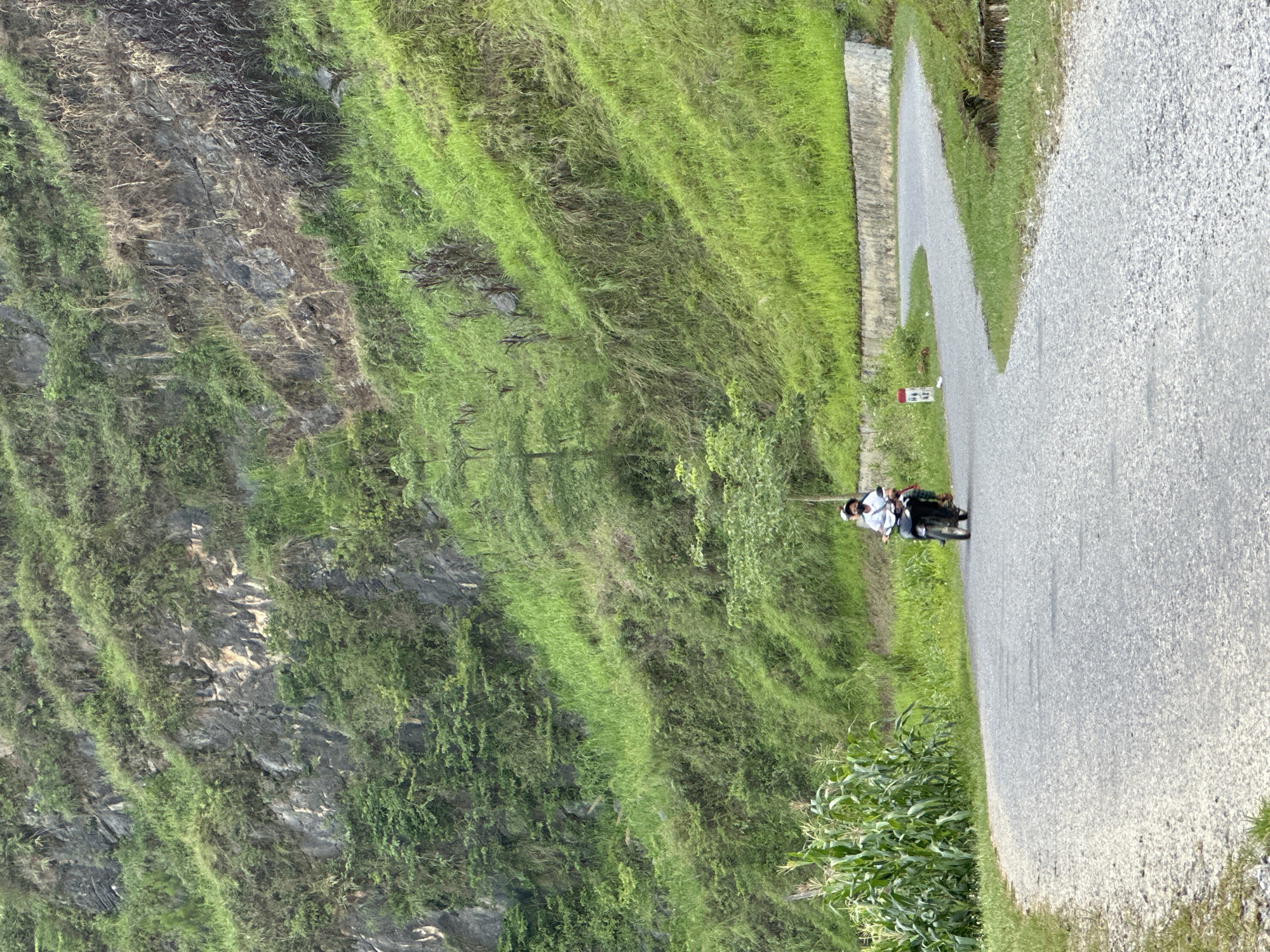 The image depicts a rural scene in Đường Thượng Commune, Vietnam. A paved road runs diagonally across the frame, with a person riding a motorcycle along it. The roadside is lined with green vegetation and a field of corn. To the left, a steep, rocky hillside covered in vegetation dominates the landscape.
