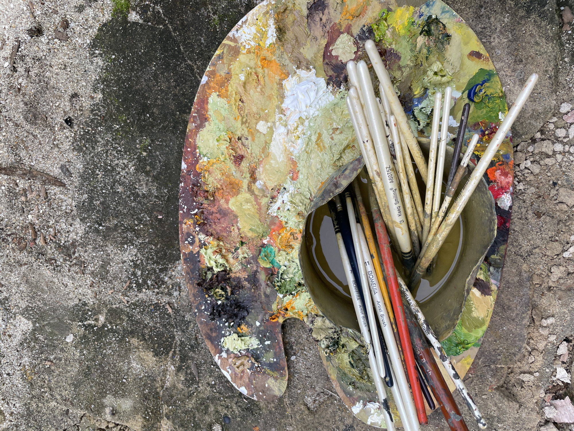 A well-used wooden artist's palette, covered in thick, impasto oil paints in shades of yellow, green, white, orange, brown, and black, rests on a rough, textured concrete surface. A collection of paintbrushes of varying sizes and colors, with some dipped in a murky, yellowish liquid within a metallic container, are placed on top of the palette. The background shows a coarse, sandy ground mixed with small stones and dried leaves, suggesting an outdoor setting. The image is taken from a high-angle perspective. There is no indication of specific location in the city of Ado, Nigeria within the image.