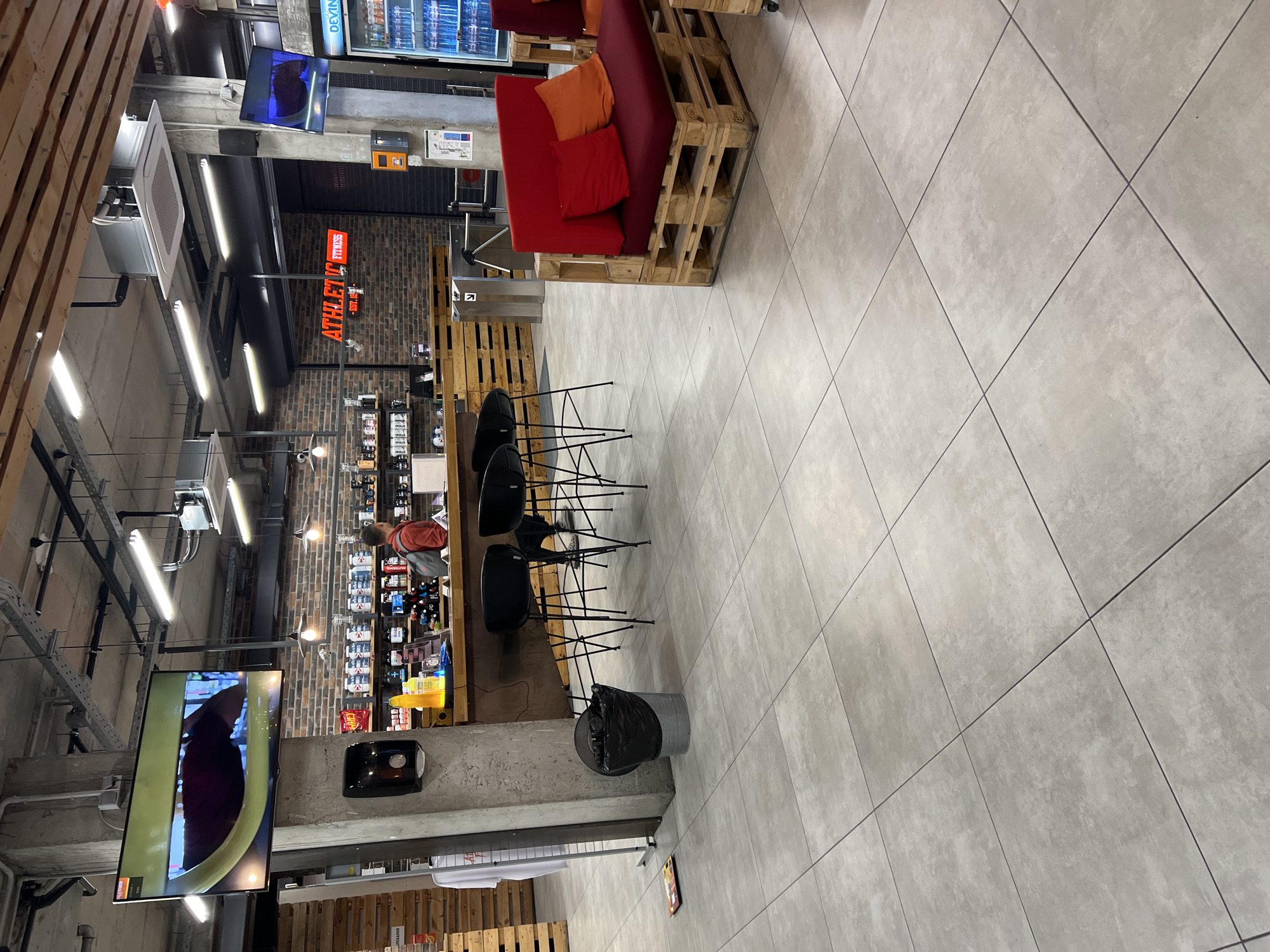 The image captures the interior of a shop, identifiable by the "ATHLETIC FITNESS" sign, suggesting it may be a fitness-related store in Sofia, Bulgaria. The store has a modern industrial design, with visible ductwork, metal beams, and a concrete structure visible near a TV screen displaying a video. Merchandise is displayed on shelves. A person in an orange top can be seen browsing. A pallet-style seating area with red cushions provides a casual seating area. The flooring is tiled in light grey, and a cylindrical trash can stands nearby. The presence of a bar counter with high stools further suggests it might be a fitness center or a store with a lounge area.