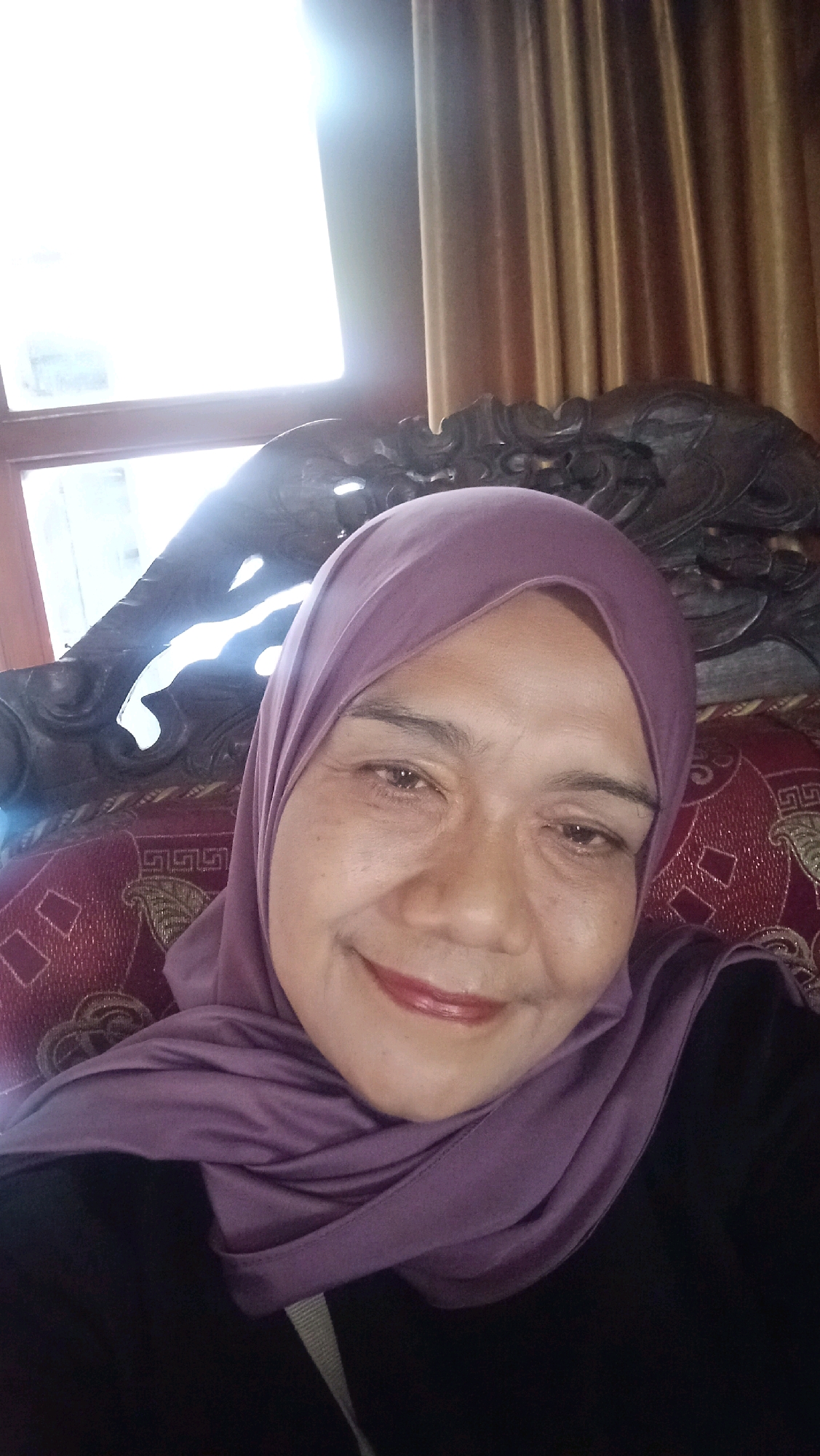 An Indonesian woman wearing a purple hijab and a black shirt smiles into the camera. She is seated on a patterned red sofa, with a dark, ornate wooden chair behind her. The background consists of a window with bright light streaming through and a tan curtain. The image was captured in the city of Kebonagung, Indonesia.