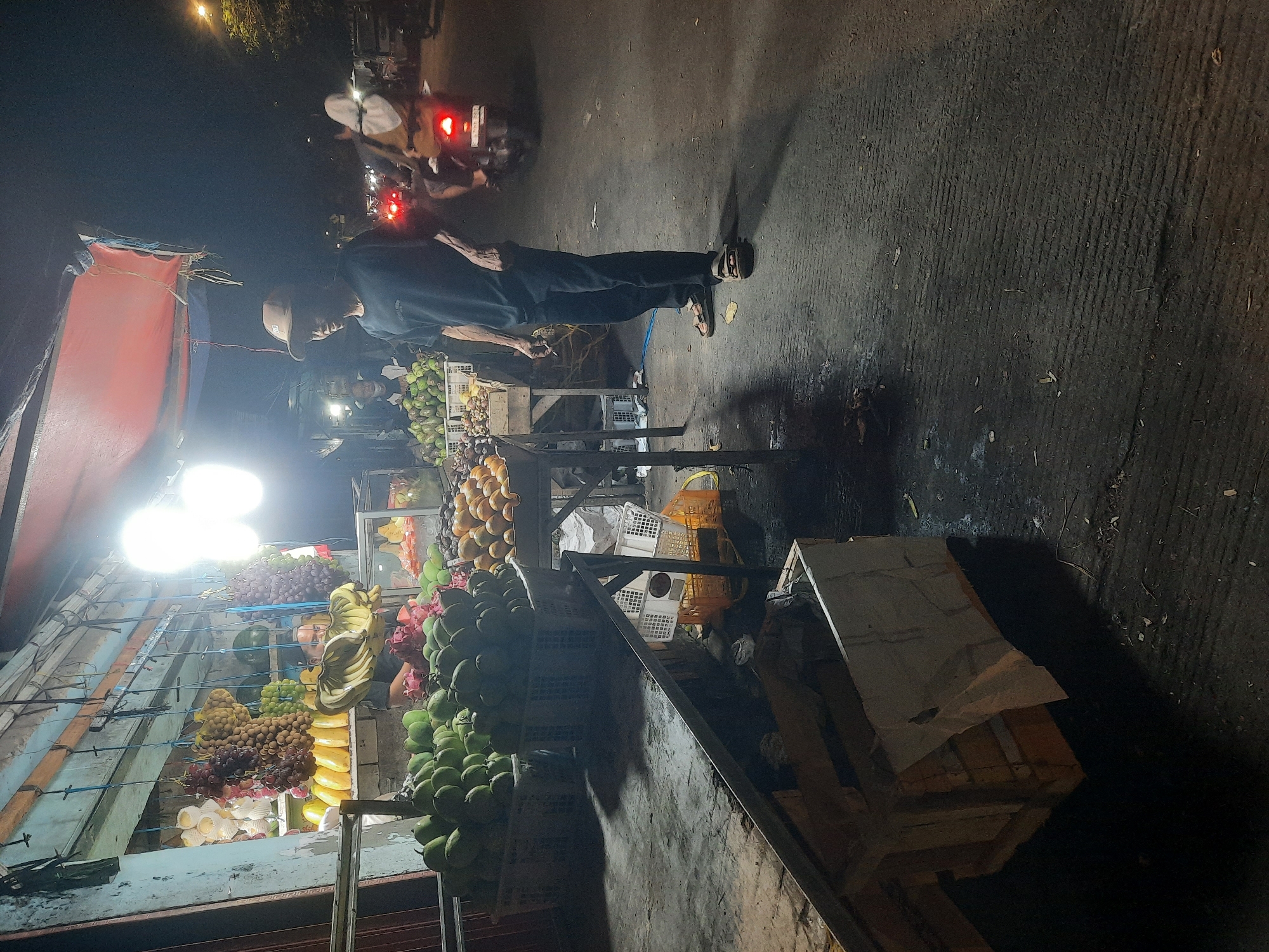The image depicts a nighttime scene at a fruit stand in North Jakarta, Indonesia. A vendor, wearing a dark t-shirt and black pants, stands near a display of fruits, including green avocados and dragon fruit. He is wearing a light-colored baseball cap and sandals and appears to be smoking. The fruit stand is illuminated by bright lights, casting shadows on the pavement. In the background, motorcycles with illuminated taillights are seen on the street, suggesting the presence of traffic. The general environment appears to be an urban street setting.