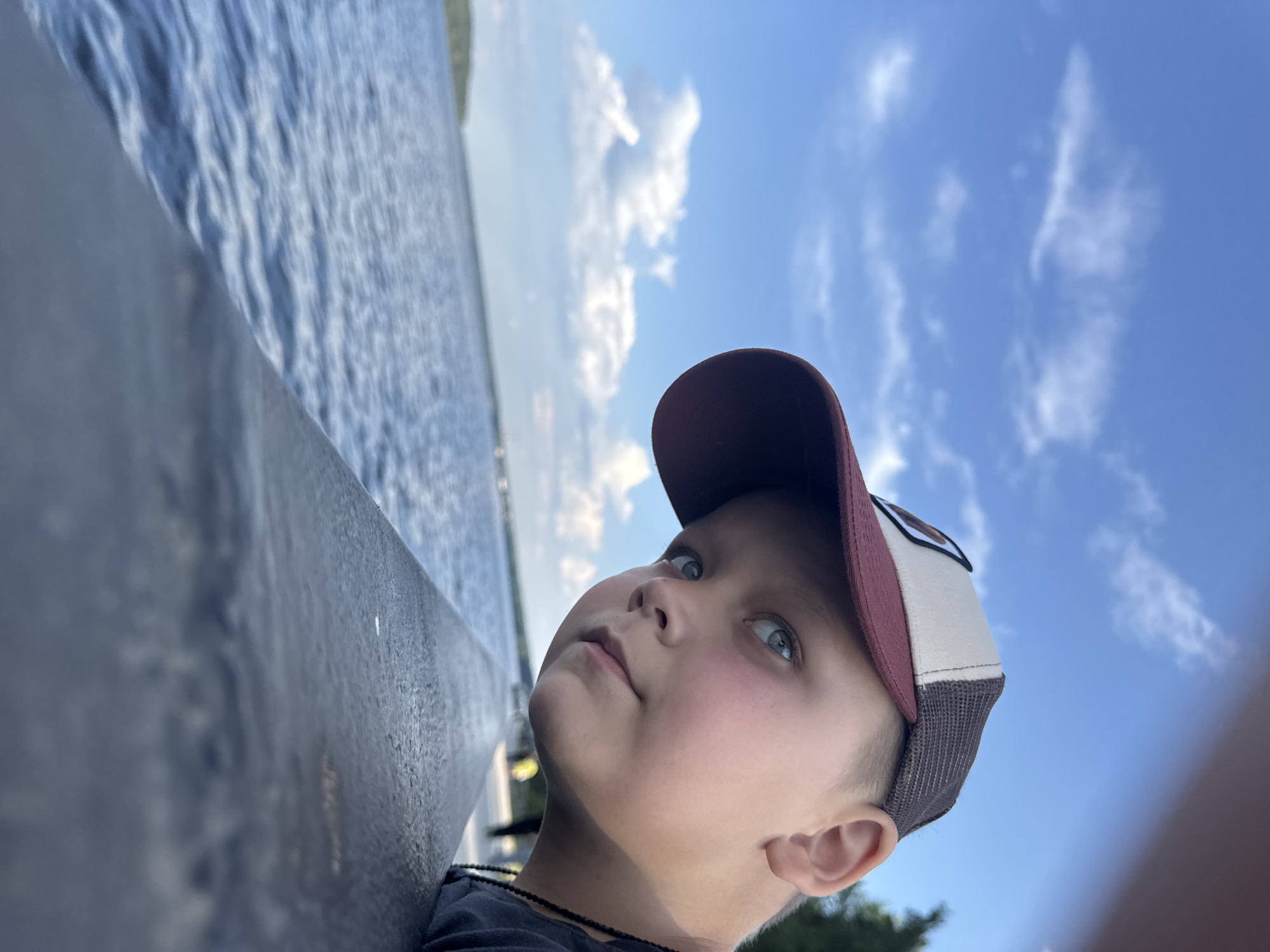 A young boy with blue eyes and wearing a baseball cap leans over a railing, looking out at a body of water. The sky above is blue with scattered white clouds. The water appears calm, with gentle ripples. The railing is dark and textured. In the background, trees and a distant shoreline are visible. The image is taken outdoors, possibly at a park or waterfront area in the city of Chaikovsky, Russia. The boy is the primary subject, and his gaze suggests contemplation or observation of the surroundings.