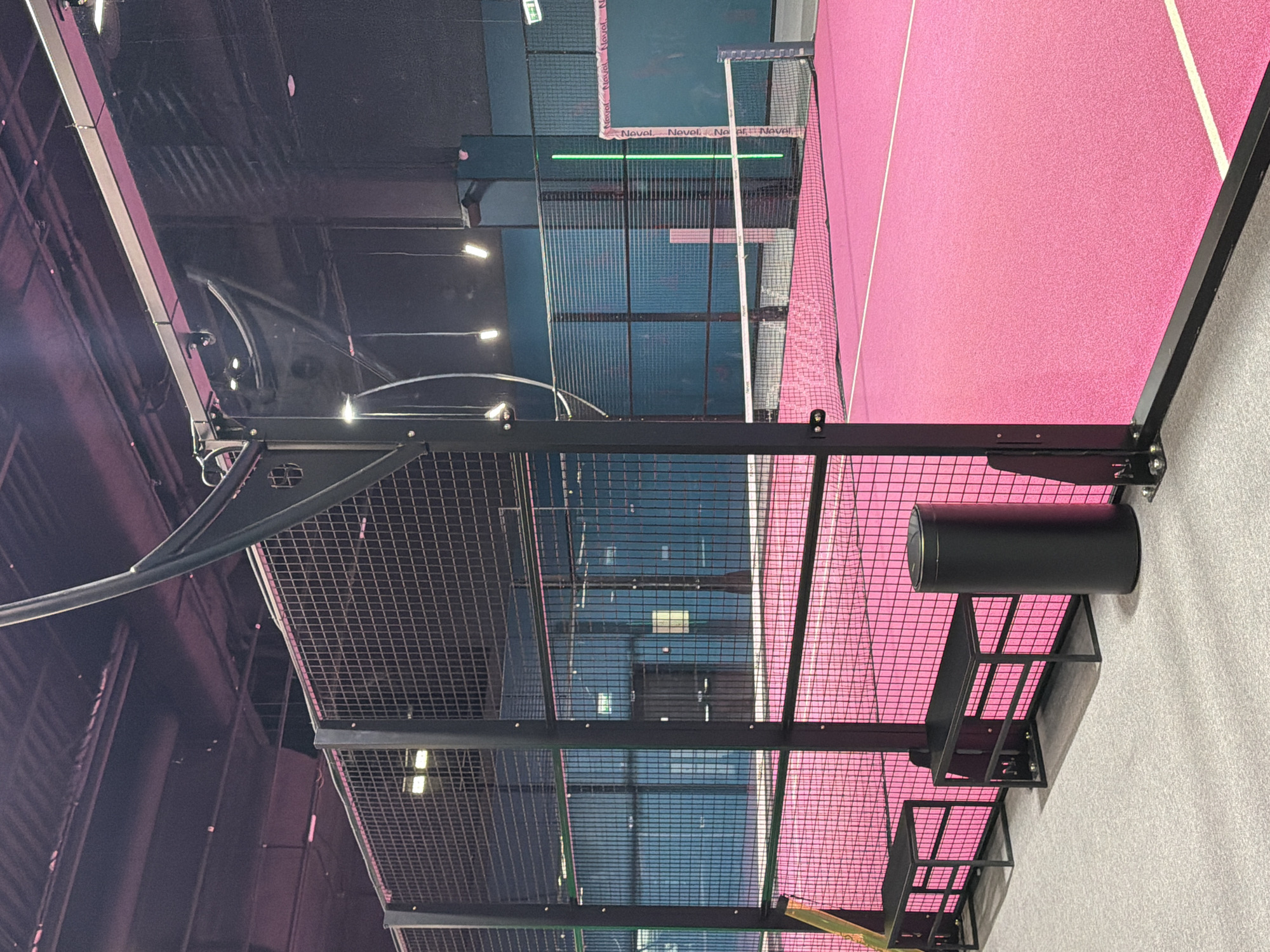 The image depicts the interior of a padel tennis court, likely indoors, with pink flooring and blue walls made of glass and wire mesh. The court is enclosed by a metal frame structure. A black cylindrical object, possibly a ventilation duct, is mounted to the wall. The location is plausibly Moscow, Russia, as the image was extracted from a larger set of images related to that city. No people are visible.