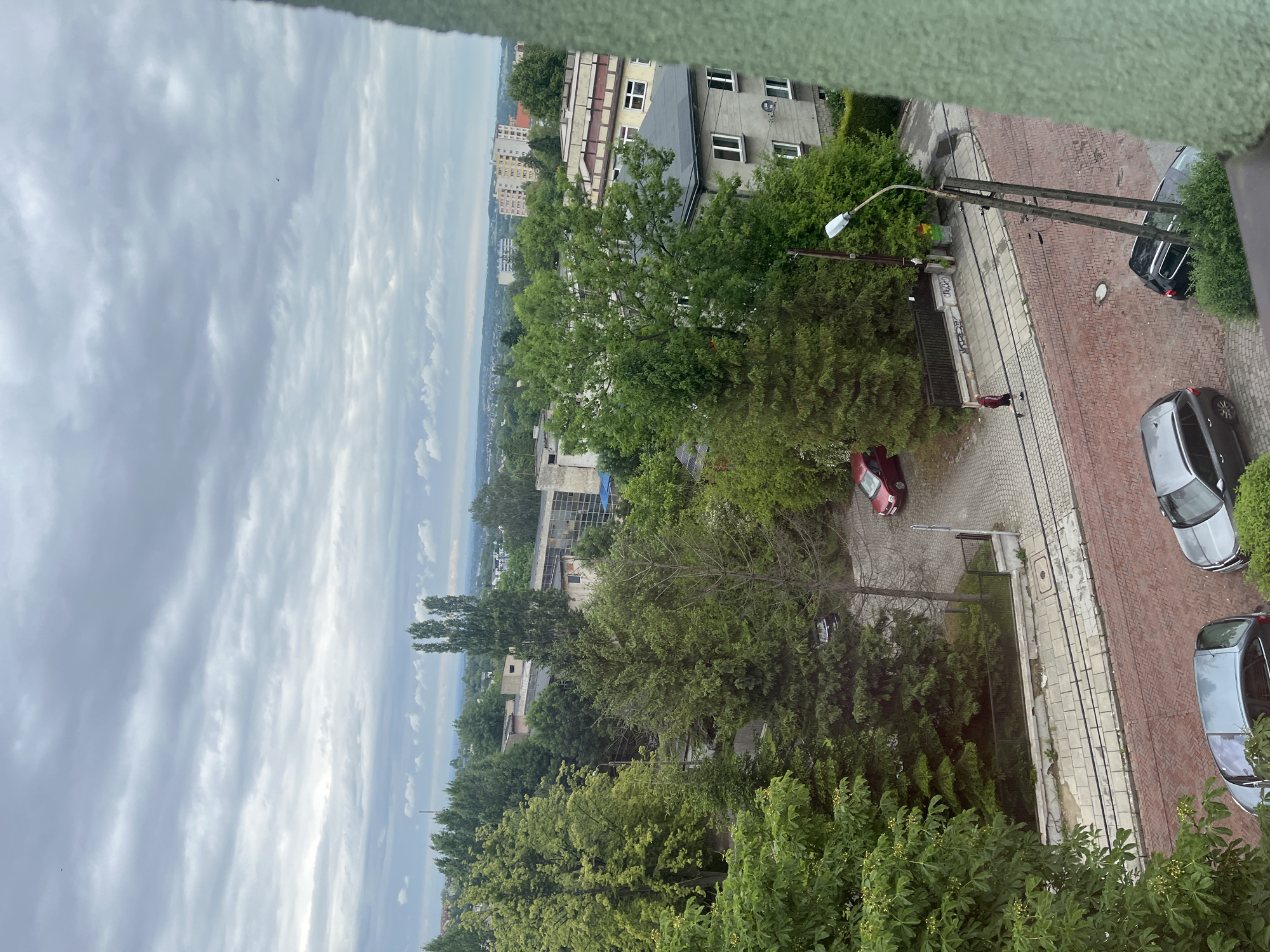 This is a high-angle, eye-level medium shot of a street scene in an urban environment under a cloudy sky. In the foreground, a green, textured wall partially obstructs the view on the right. Below this, a red brick pavement runs diagonally across the image, with two parked cars visible: a silver SUV on the left and a black SUV on the right. A light pole with wires stands in the middle of the paved area. Further back, a sidewalk is visible, with a person walking from left to right. A red car is parked on the sidewalk. Behind the sidewalk, a fence separates it from a tree-lined area and buildings. Large green trees dominate the mid-ground, with various buildings of different sizes and architectural styles visible beyond them, extending into the background. The sky is overcast with dense grey clouds, with some lighter patches indicating breaks in the cloud cover. The overall impression is a somewhat overcast day in a residential or mixed-use urban area.
