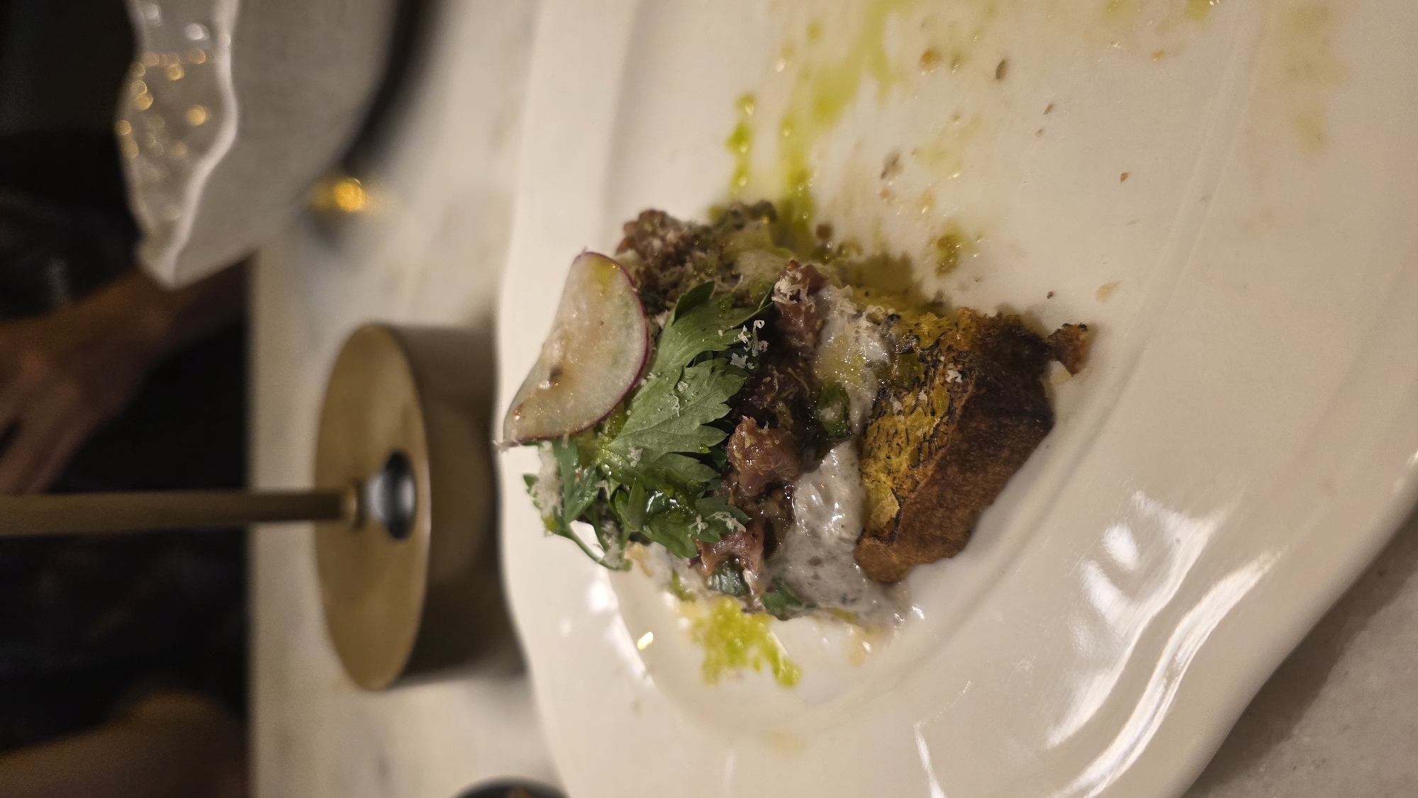 The image depicts a partially consumed dish on a white plate. The remaining portion includes a piece of toasted bread topped with what appears to be meat, herbs (likely parsley), a radish slice, and a creamy sauce. Traces of olive oil are visible on the plate. A metallic, cylindrical object with a stick extending from it is placed to the left of the plate. The scene appears to be indoors, possibly a restaurant setting. The location, Fethiye, Turkey, is not discernible from the image itself.