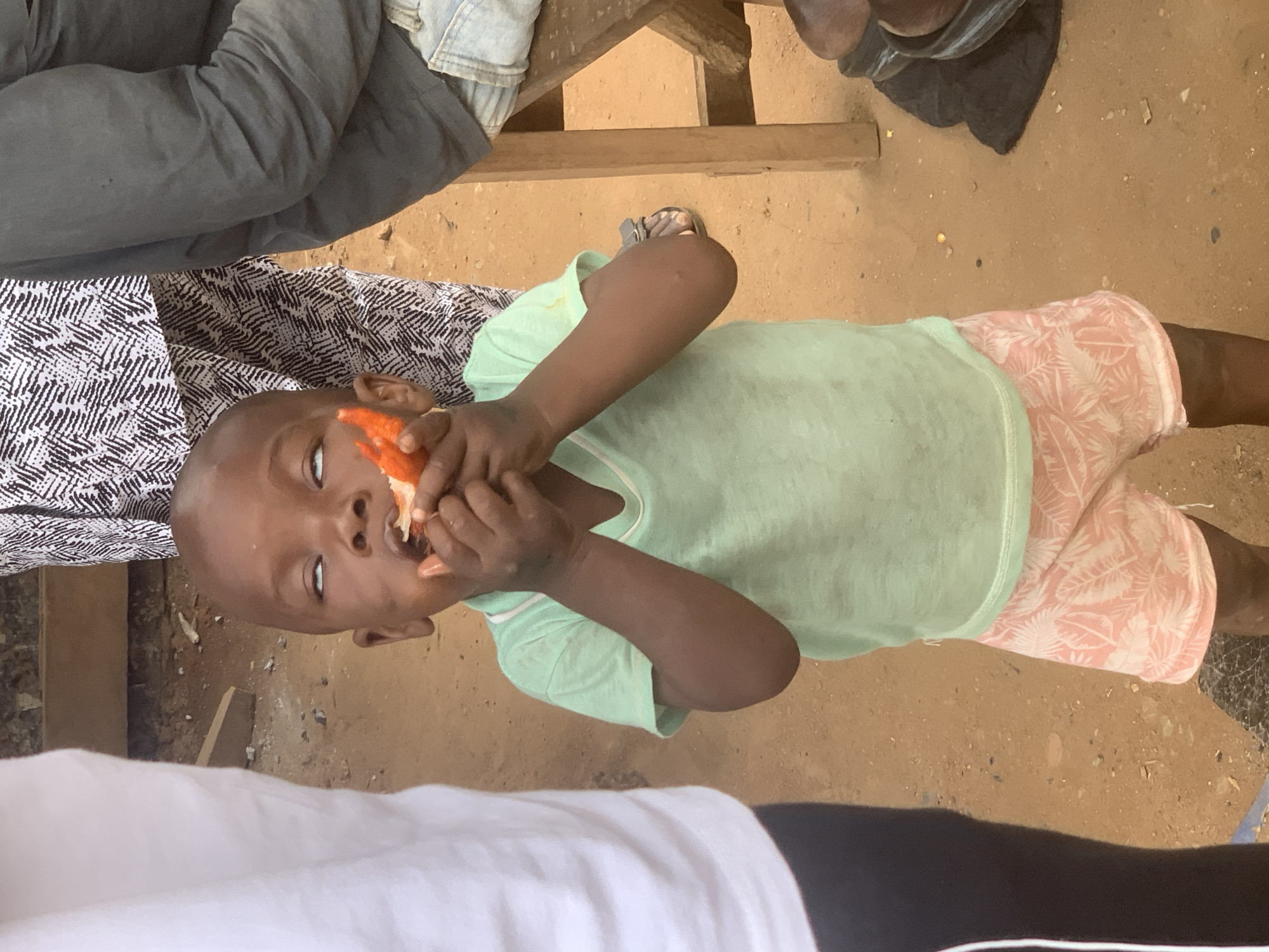 A young child is eating a piece of food with their hands while standing in a dirt area. The child is wearing a light green t-shirt and pink and white patterned shorts. Their eyes are rolled back. In the background, there are people and a wooden bench. The scene appears to be outdoors, possibly in a market or a street in Shagamu, Nigeria.